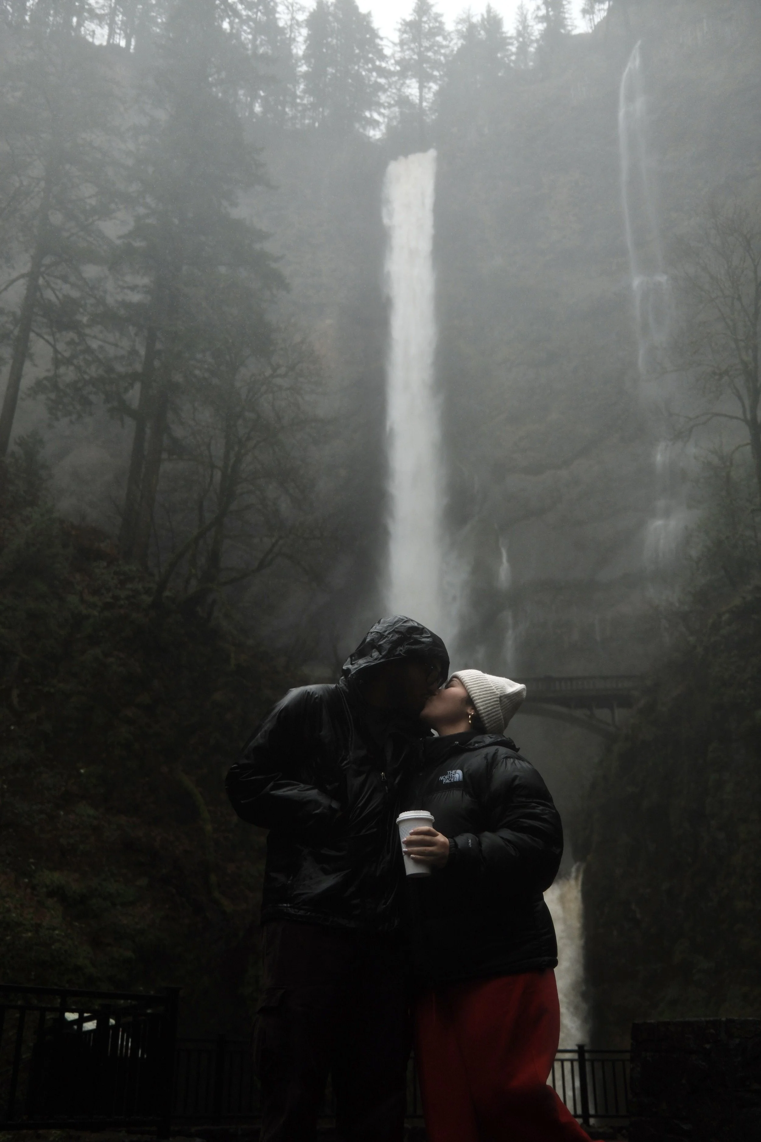 A couple kissing in front of a waterfall, with trees and mist in the background, while holding coffee cups.