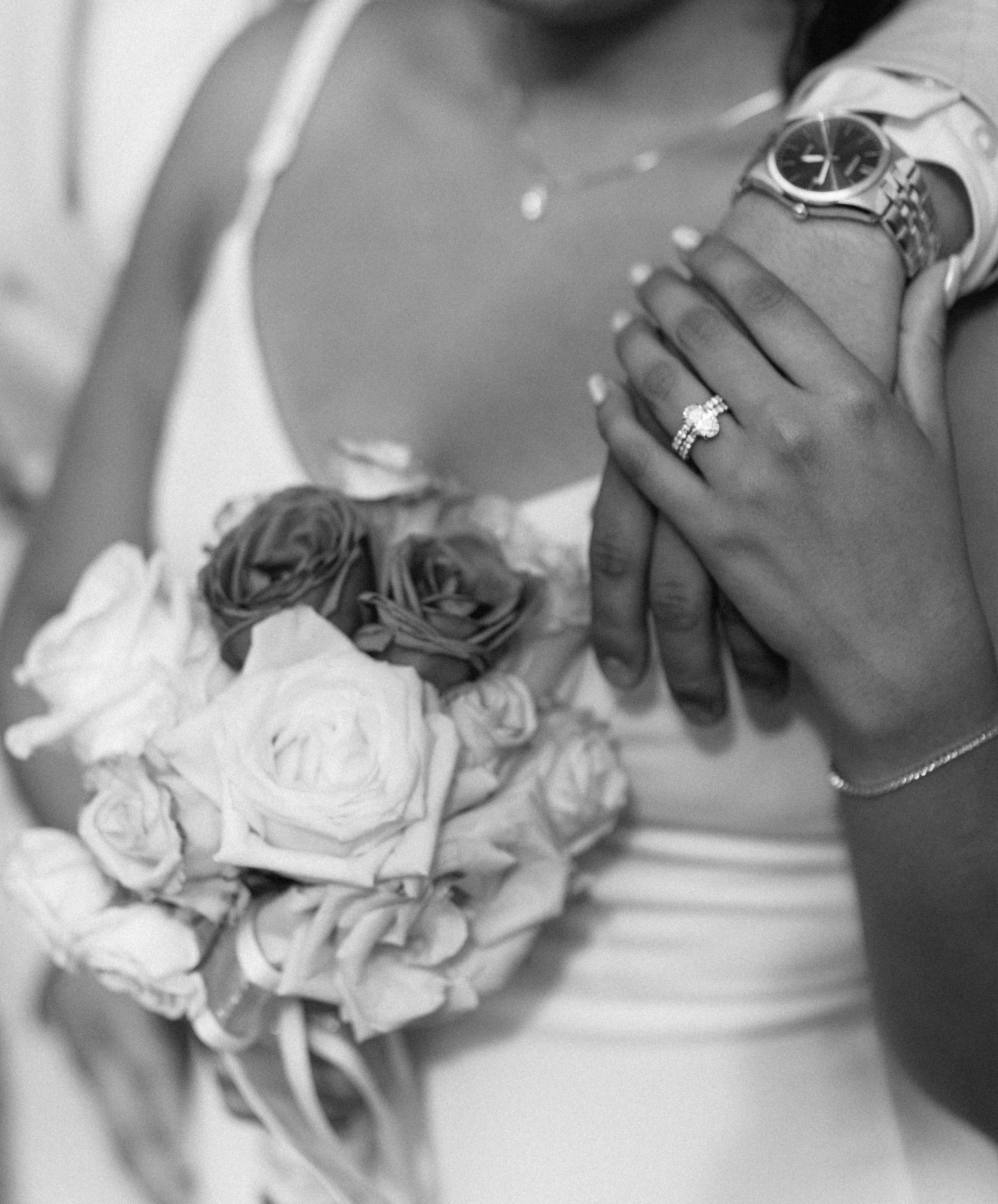 Close-up of a woman's hand showing a wedding ring, a wristwatch, and a bracelet. The woman is holding a bouquet of roses, and part of her dress is visible.