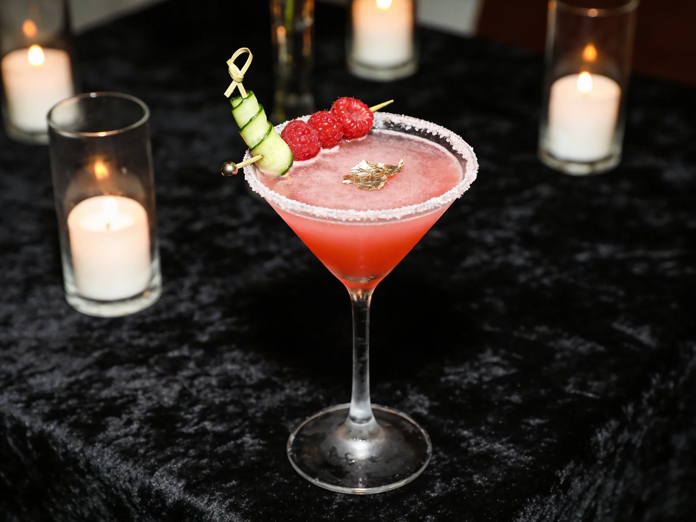 Pink cocktail in a martini glass with a salted rim, garnished with cucumber slices, raspberries, and an edible gold leaf, on a black velvet table surrounded by candles.
