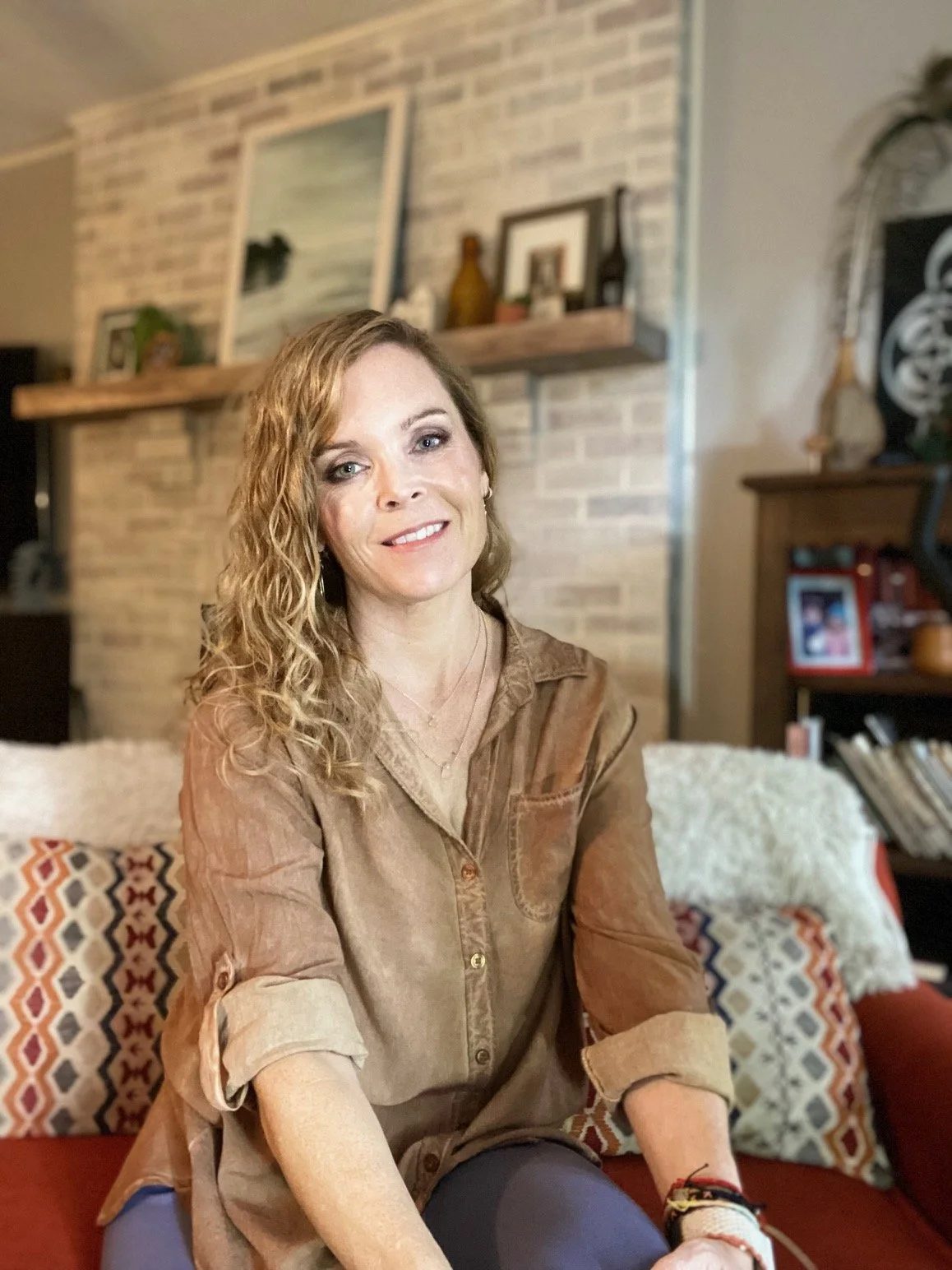 A woman with curly blonde hair sitting on a red couch in a living room with a brick wall background, decorated with framed pictures, a shelf with bottles and ornaments, and patterned pillows.