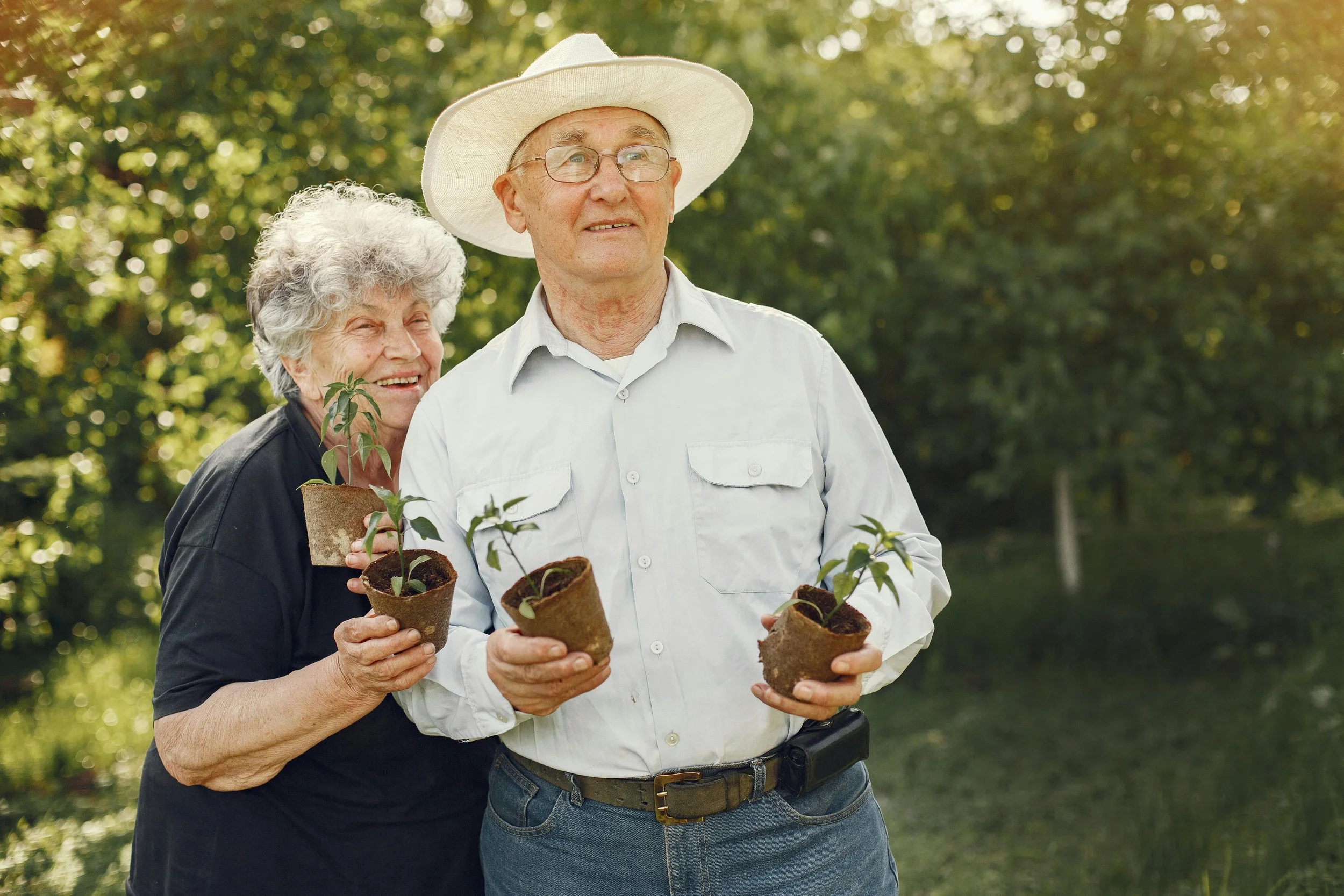 An elderly man and woman outdoors holding small potted plants, with the woman smiling and the man wearing a wide-brimmed hat and glasses.