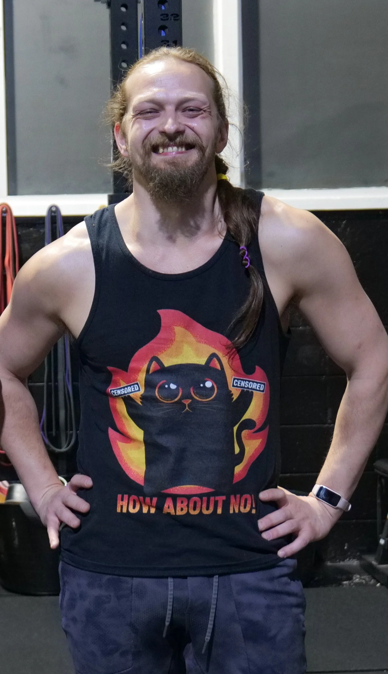 A man with long hair and a beard, smiling, wearing a black sleeveless shirt with a graphic of a black cat with large eyes and a trail of fire behind it, and the words 'HOW ABOUT NO!'