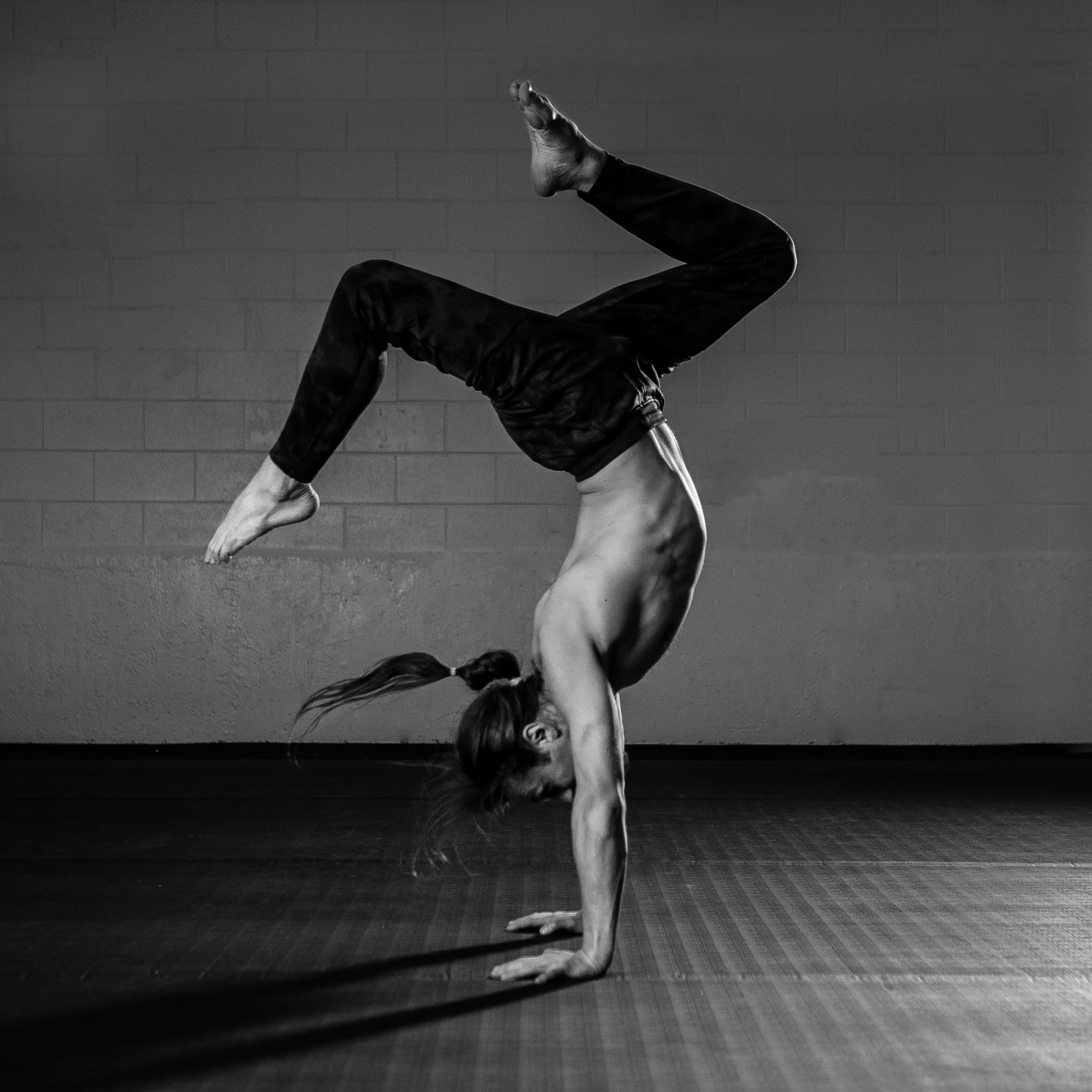 A woman performing a handstand in a yoga pose, balancing on her hands with her legs bent and one foot touching her head, in a gym setting.