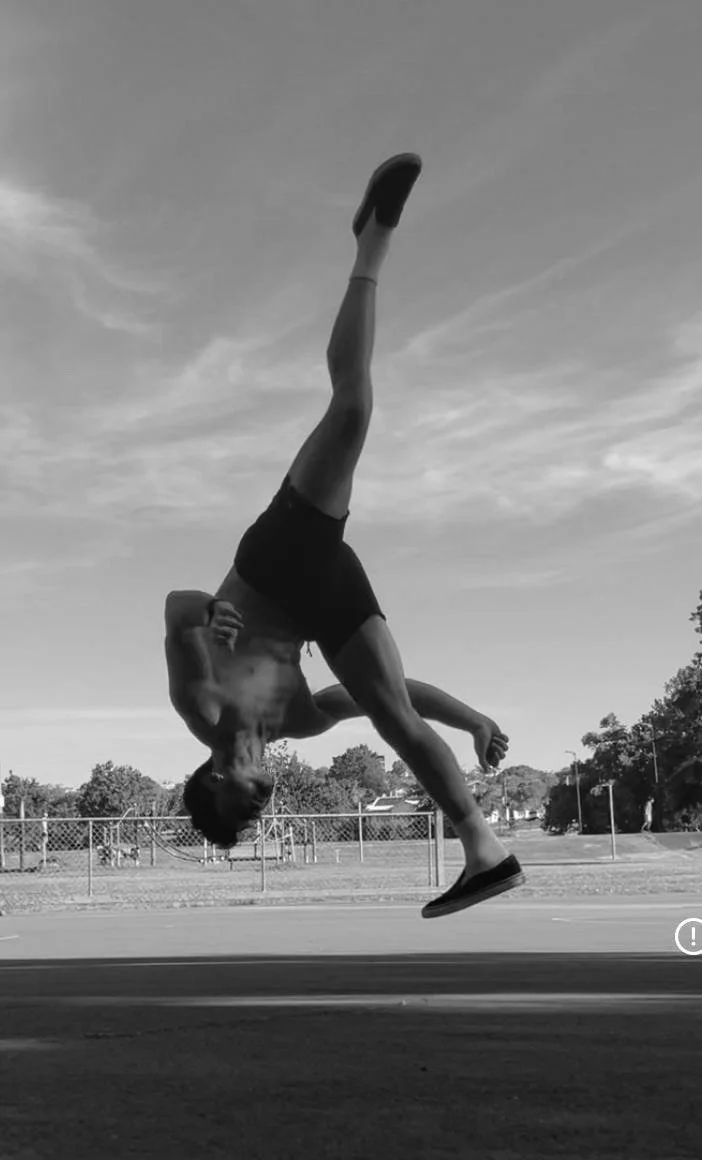 A person performing a handstand flip outdoors on a paved surface, with trees and a chain-link fence in the background, captured in black and white.