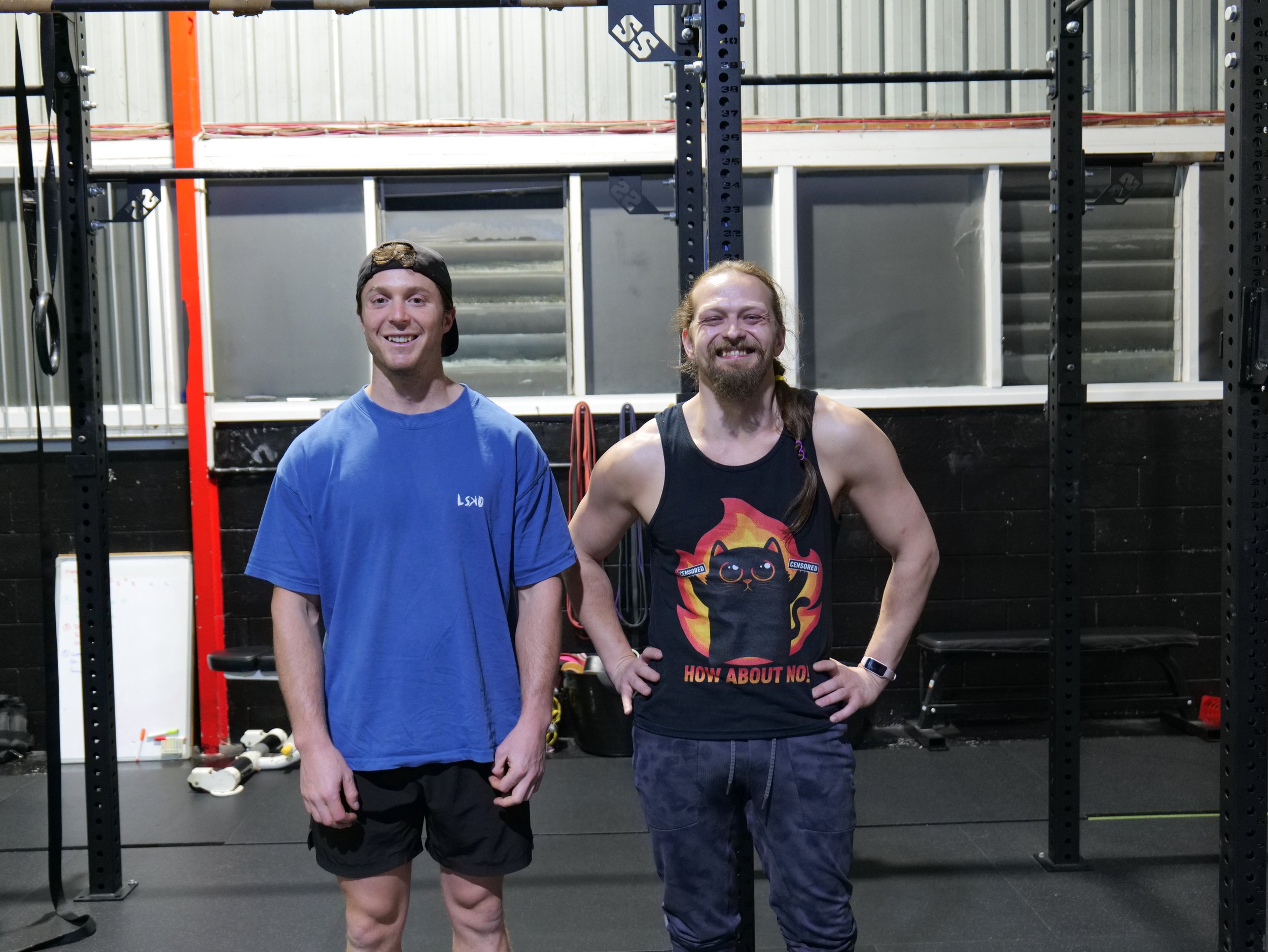 Two men standing side by side inside a gym, smiling at the camera, with black gym flooring and workout equipment in the background.