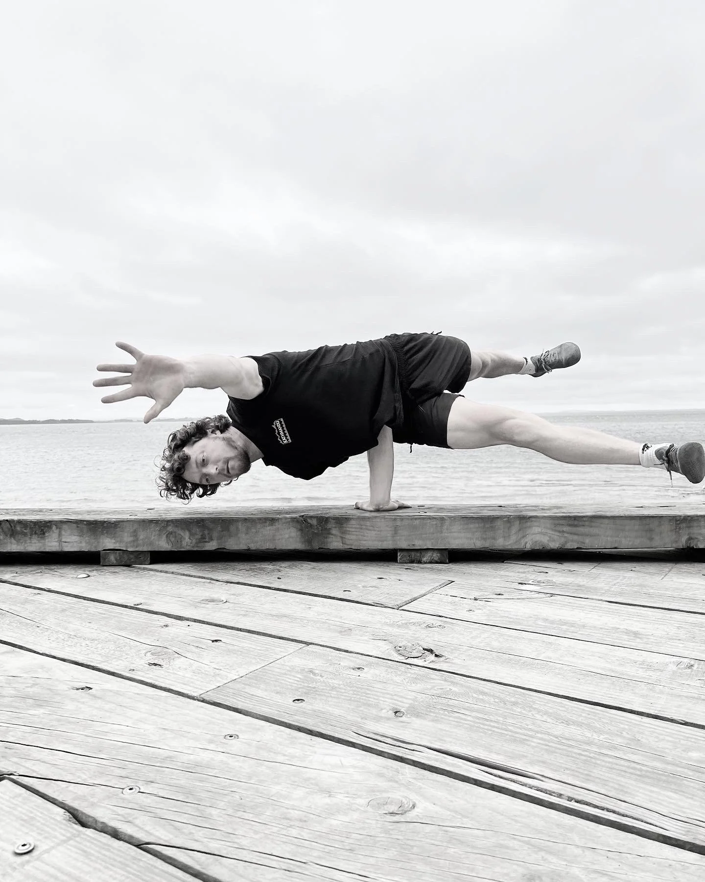 A man balancing on one hand on a wooden dock, with one arm stretched out and the other supporting his body, by the water.