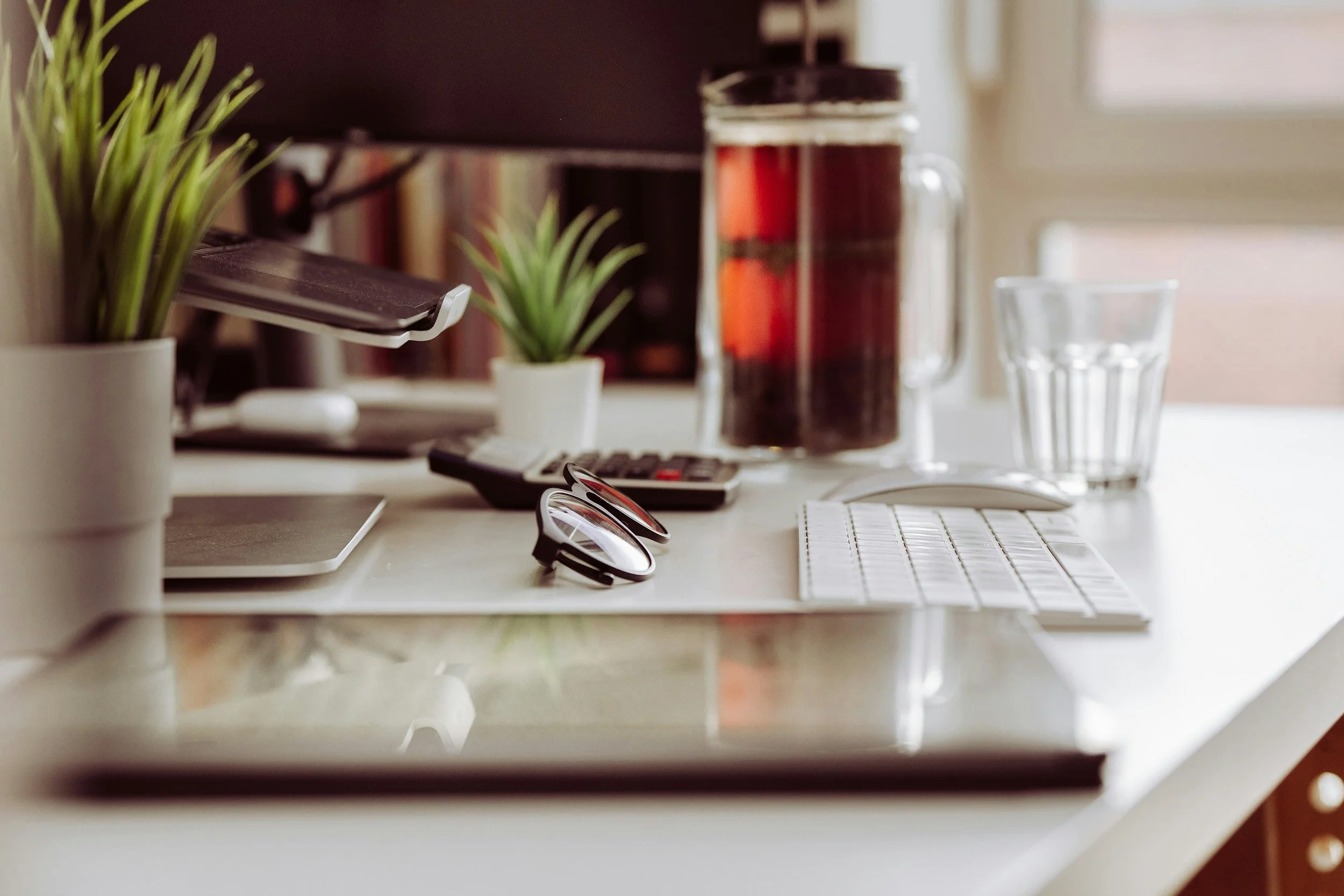 A cluttered office desk with a potted plant, eyeglasses, a calculator, a glass of water, and a large glass jar of beverage.