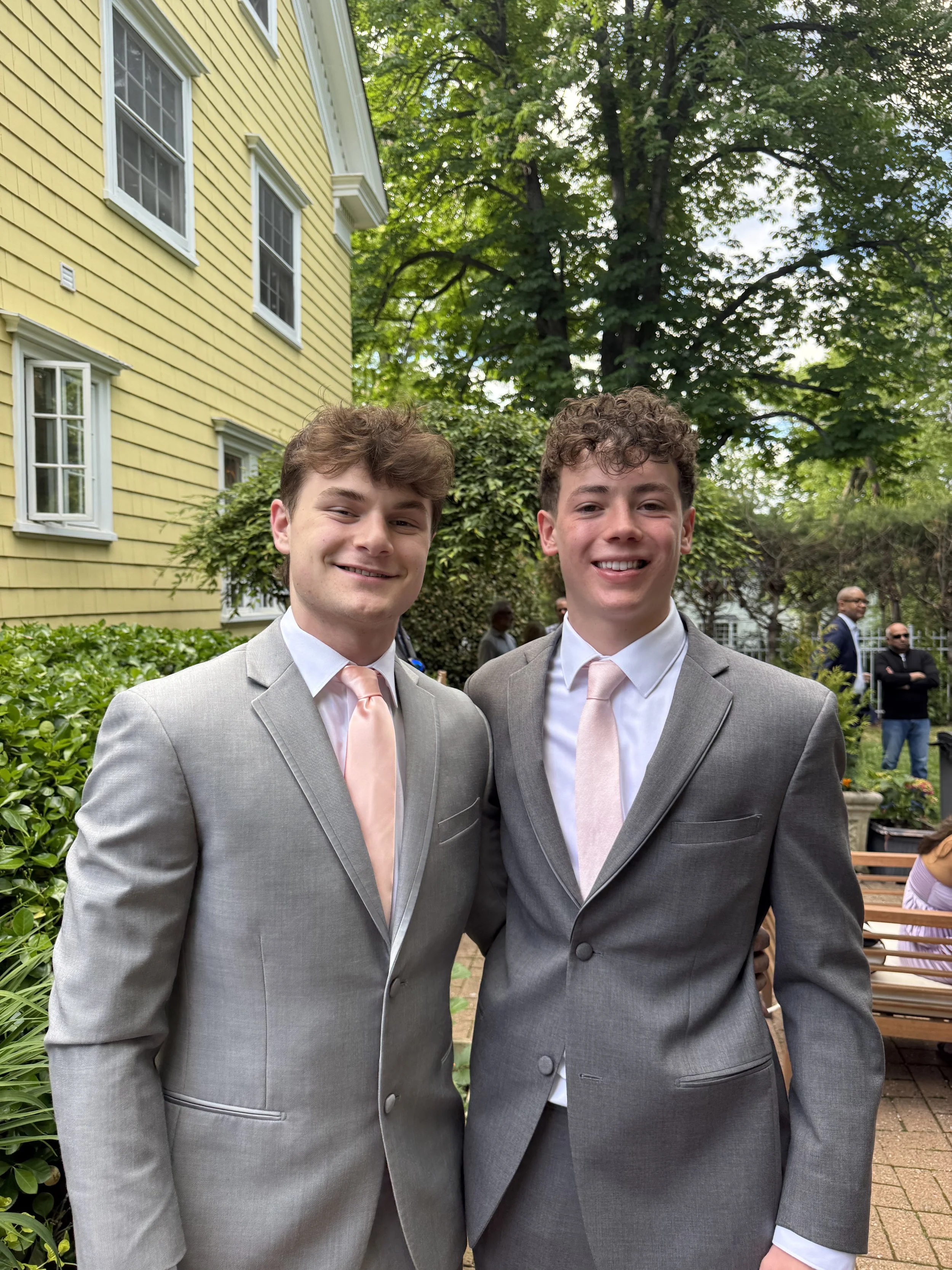 Two young men in formal suits smiling outdoors in front of a yellow house with green trees and other people in the background.