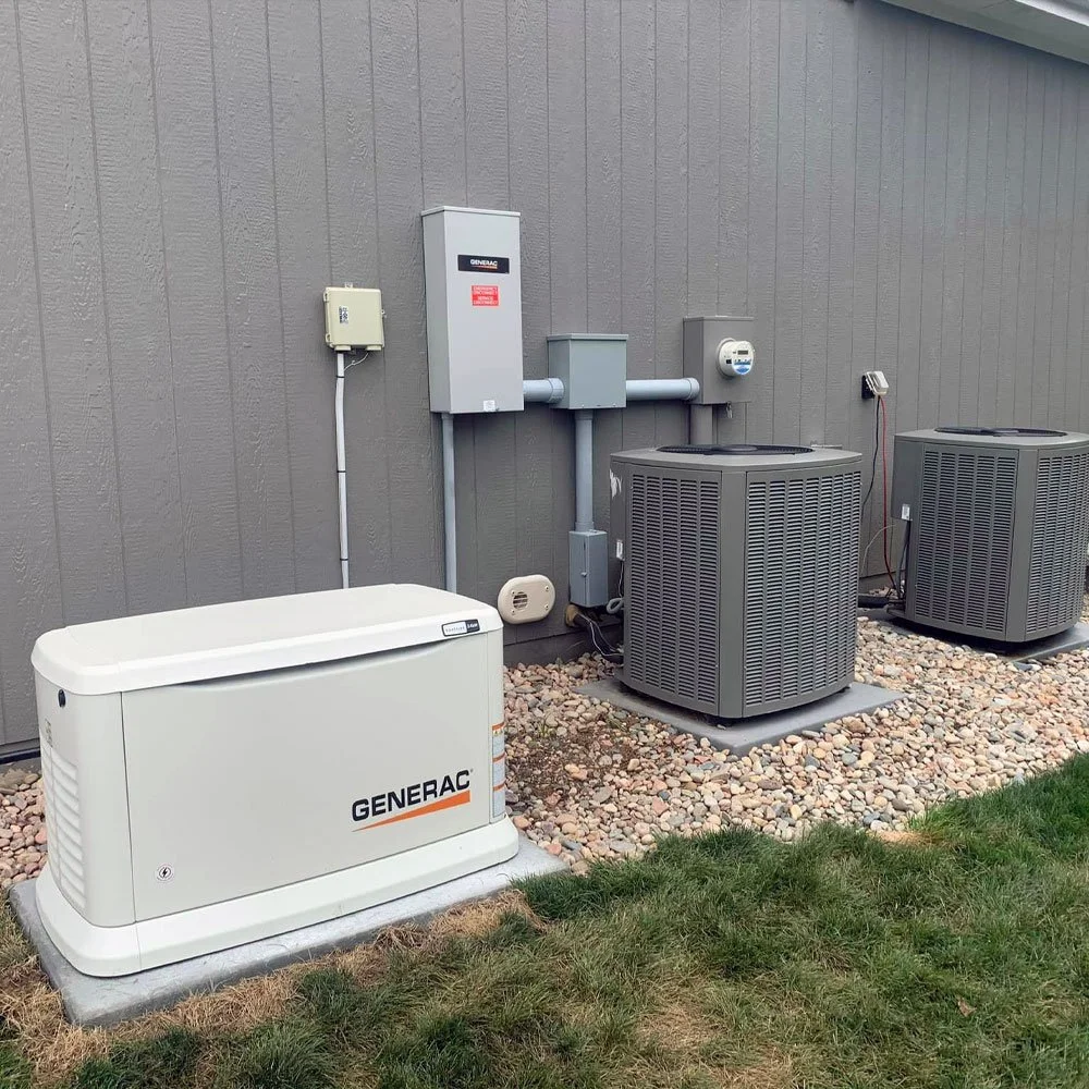 Outdoor area with climate control equipment including a Generac generator, two air conditioning units, and electrical boxes on a gray wall, with gravel and grass ground.