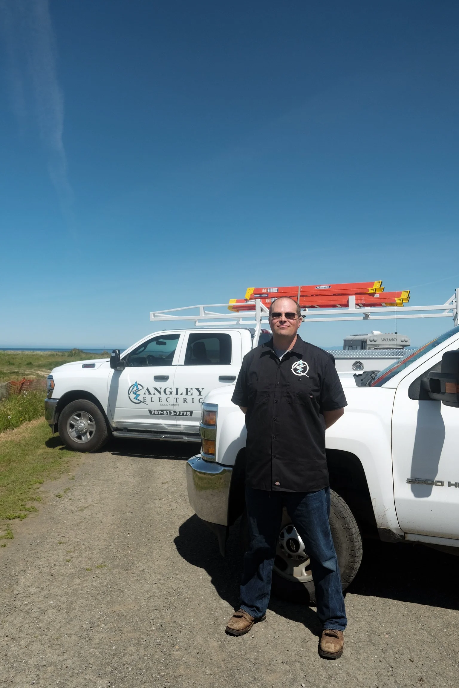 A man standing in front of a white pickup truck with the words 'Angley Electric' on the door, outdoors on a clear day, with a blue sky and green landscape in the background.