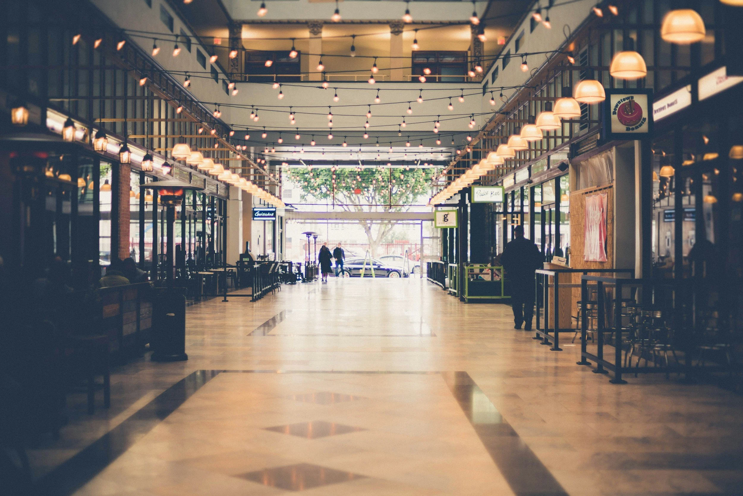 Empty indoor shopping mall corridor with string lights hanging from the ceiling and some people walking near the exit at the end of the corridor.