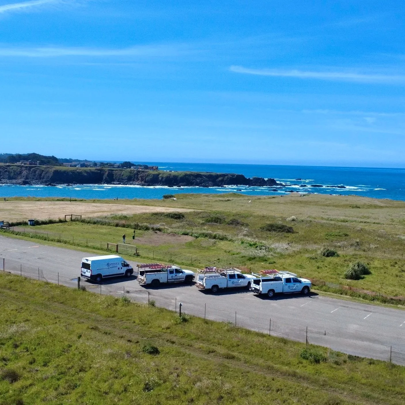A coastal landscape with a grassy area, parked utility trucks and vans, a walking person, and an ocean with rocky cliffs in the background under a partly cloudy sky.