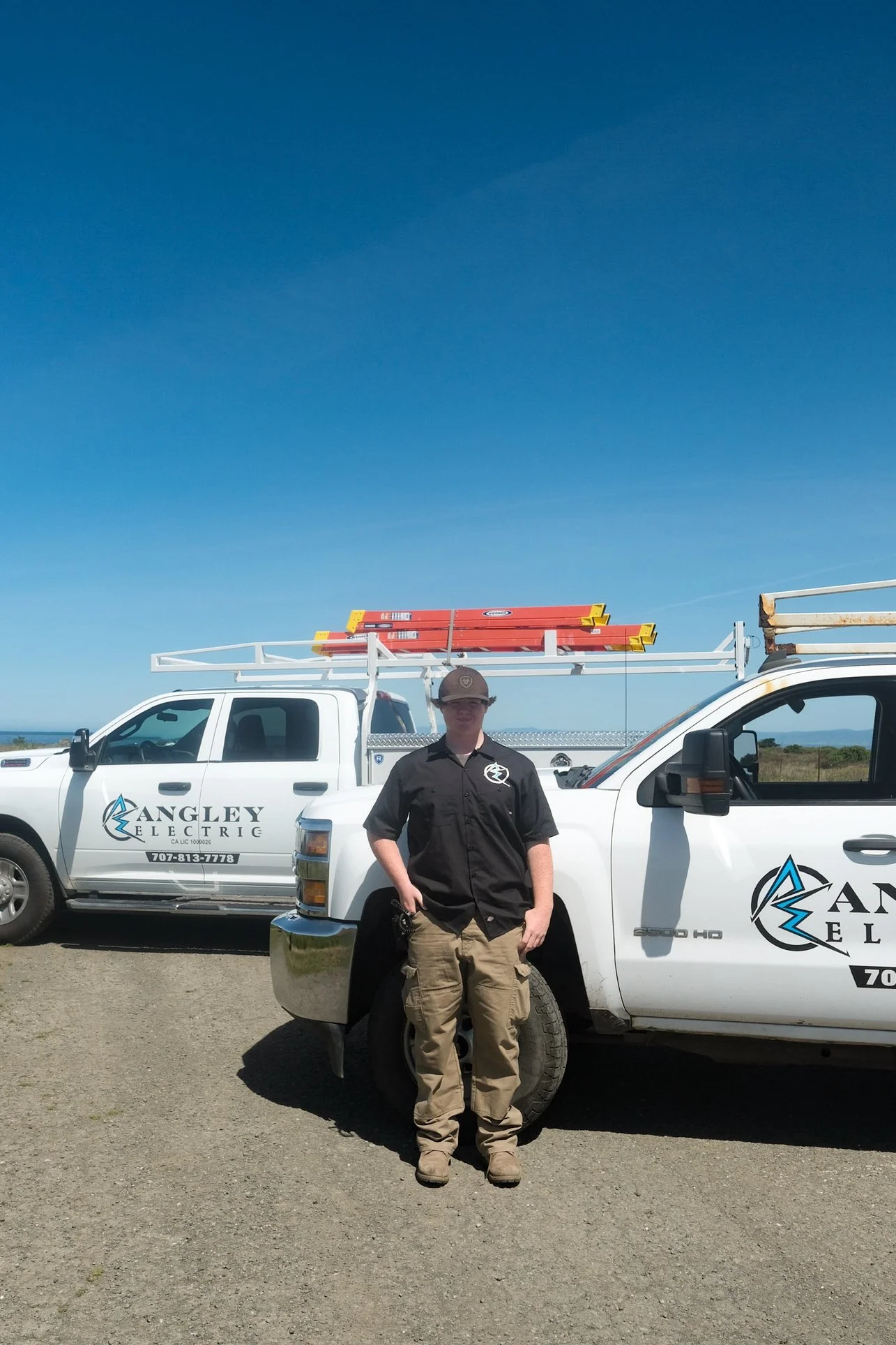 A man wearing a black work shirt, khaki pants, and a cap stands next to a white utility truck with 'Angley Electric' branding, sunny sky in the background.