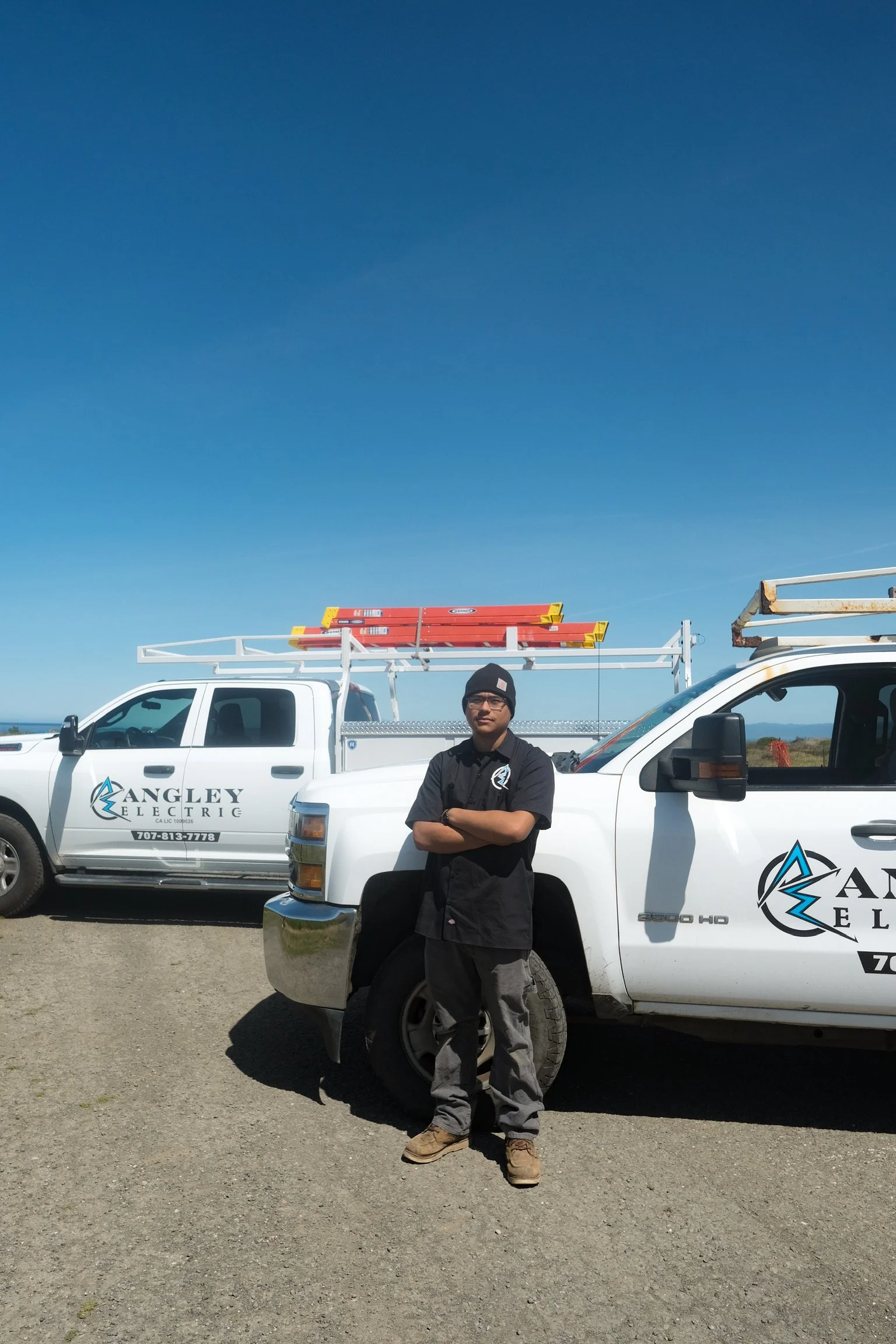 A man in work clothes standing in front of a white utility truck with a company logo, with a clear blue sky overhead and a second similar white truck nearby.