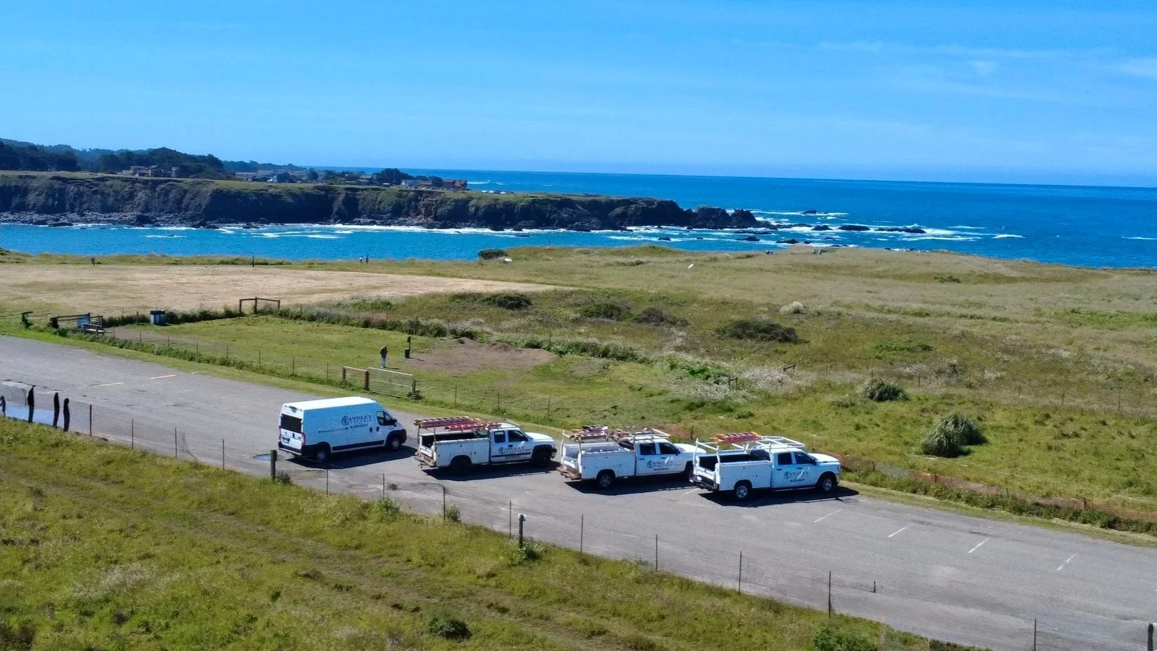 Coastal landscape with grassy field, parked white utility trucks, and ocean with cliffs in the background under a partly cloudy sky.