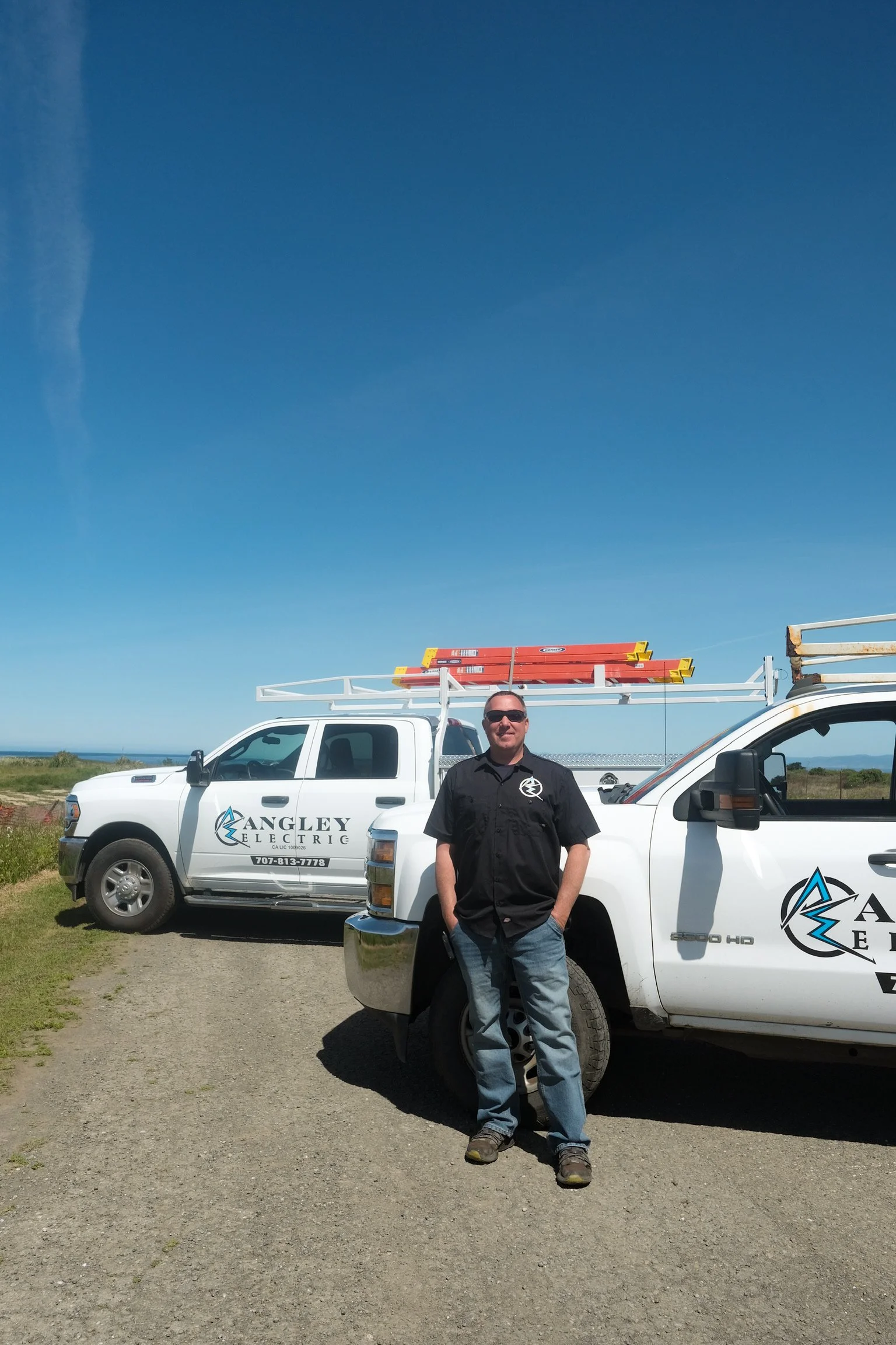 A man wearing glasses, a black shirt, and jeans, standing in front of white utility trucks with company logos and ladders on the roof, outdoors on a sunny day with a clear blue sky.