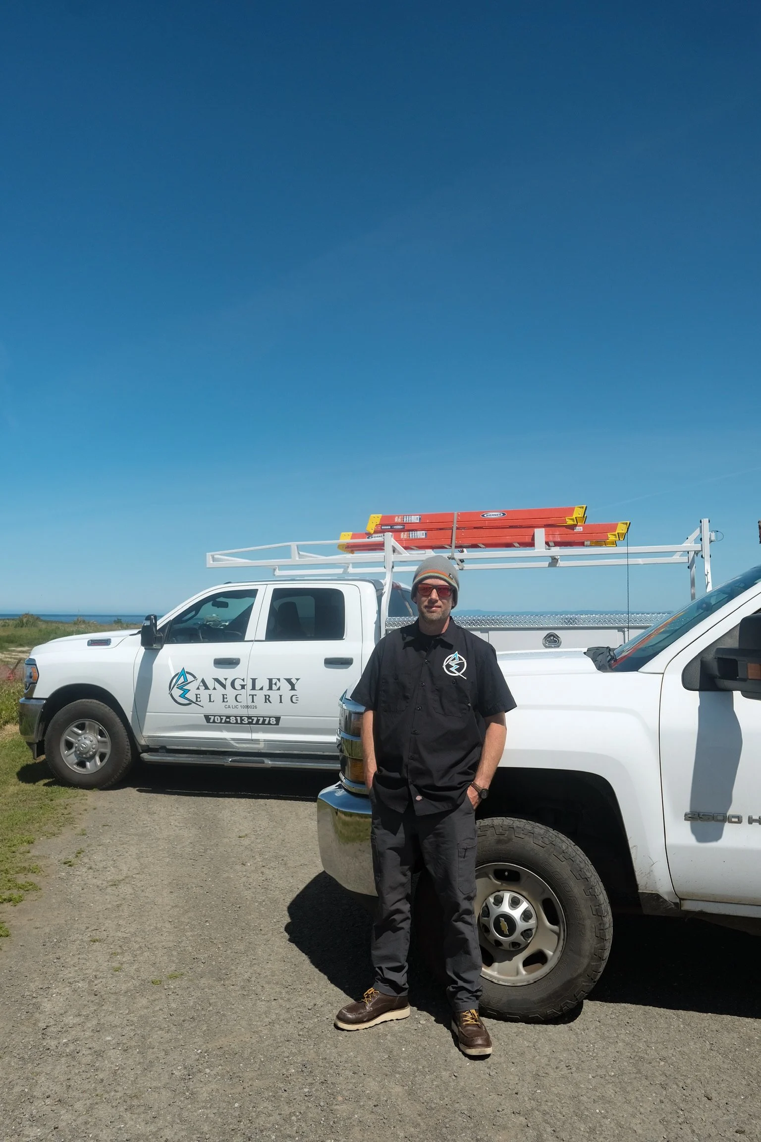 A man wearing dark work clothes and sunglasses standing in front of a white service truck with the logo 'Angley Electric' and a phone number, on a gravel road with a blue sky background.