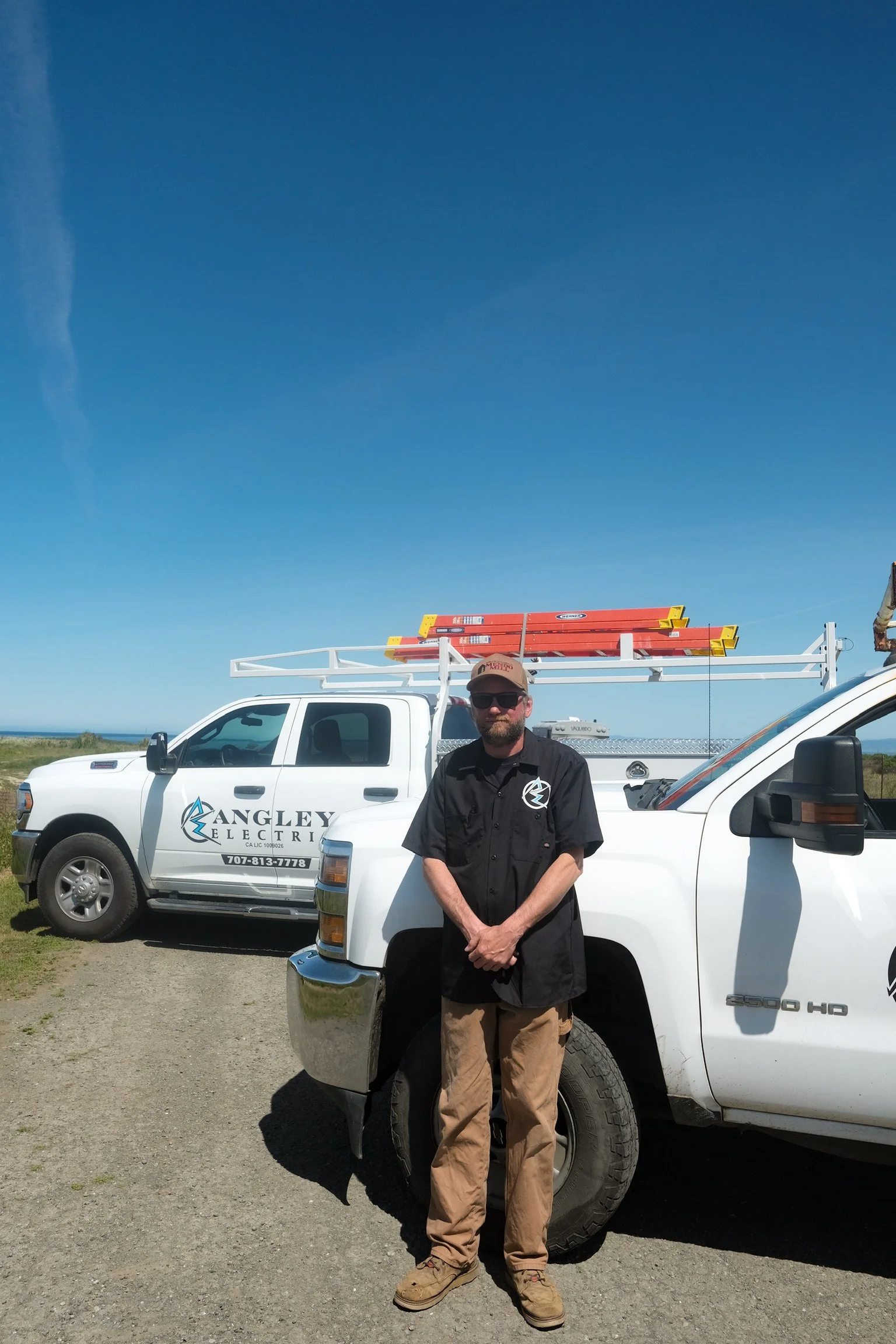 A man with a beard wearing sunglasses, a black shirt with a logo, and khaki pants, standing in front of a white utility truck with company branding 'Angley's Electric' on the door, under a clear blue sky with a yellow and red ladder on top of the tru