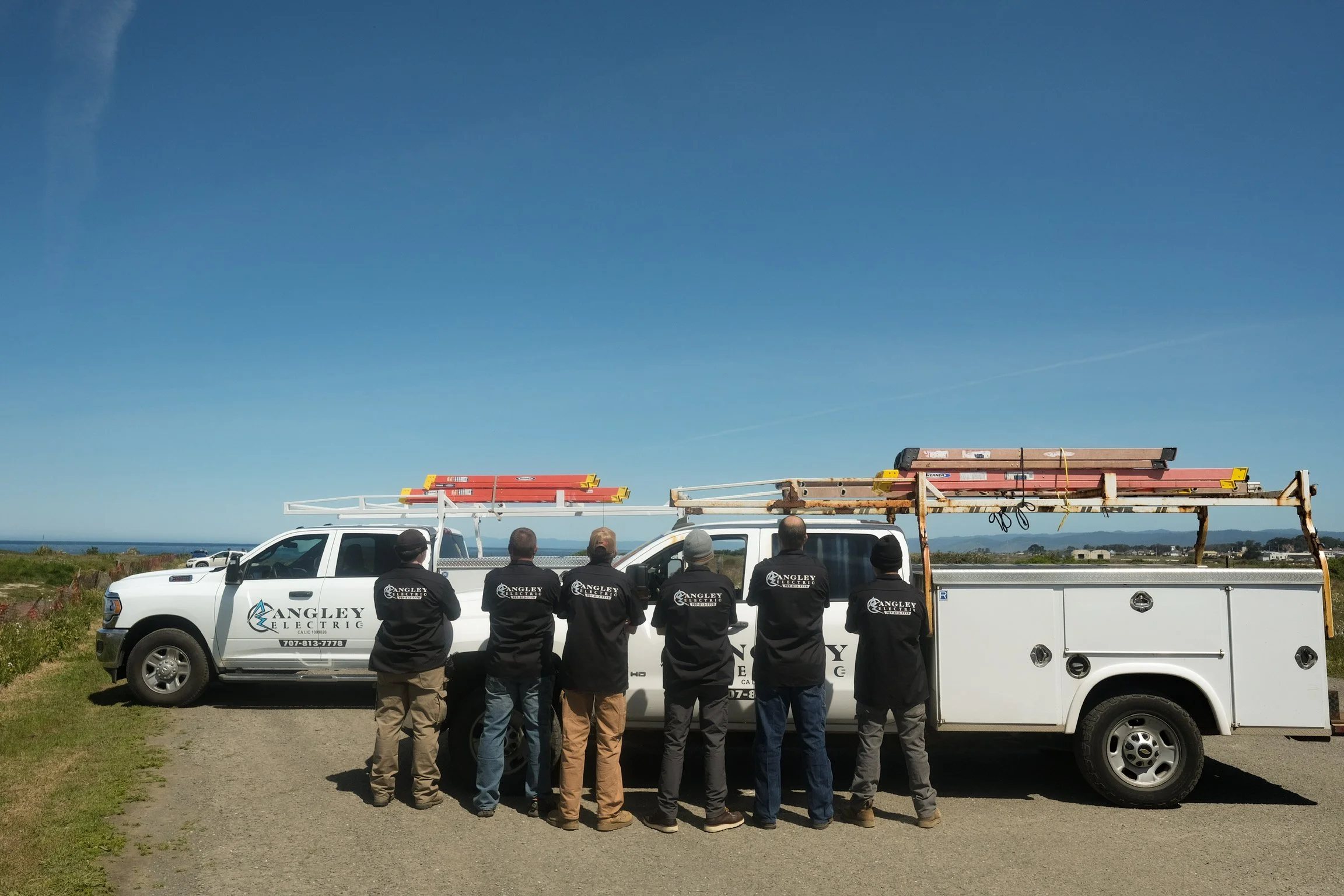 Group of workers from Angle Electric standing in front of a white utility truck with a long ladder on top, during daytime with blue sky and open landscape in the background.