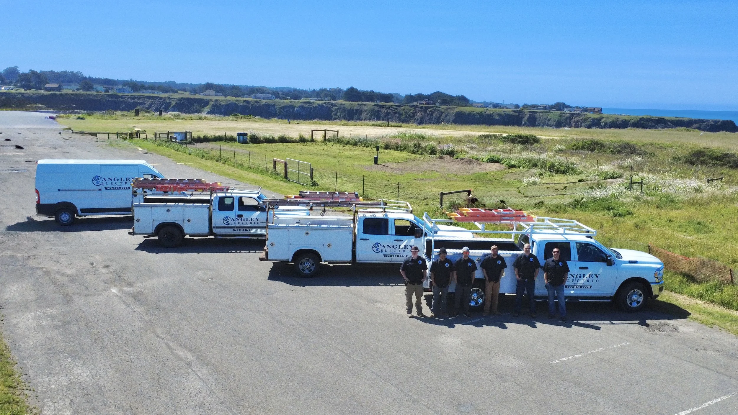 Six workers in uniform standing in front of three utility trucks labeled 'Angley Electric' in a parking area near a grassy coastal landscape with cliffs and the ocean in the background.