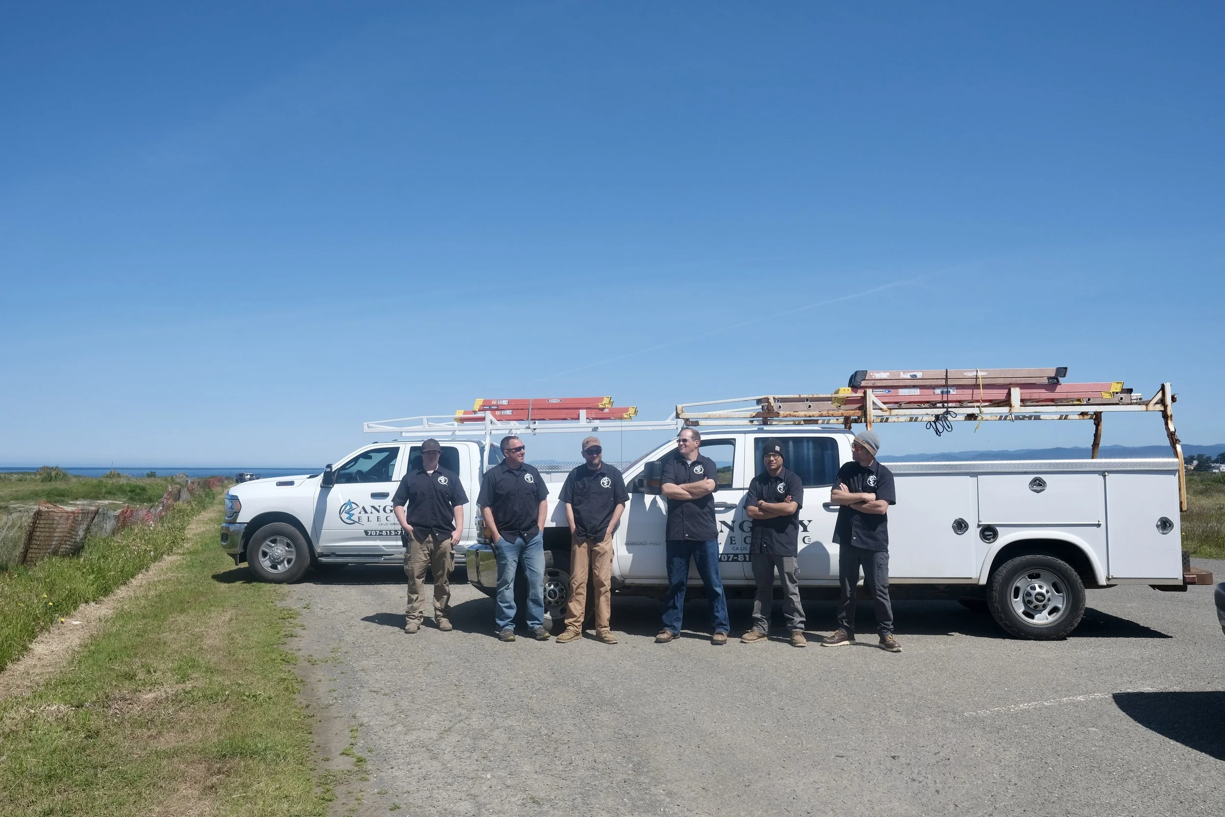Five men standing in front of a white truck on a grassy area near the coast, with a blue sky and distant mountains in the background.