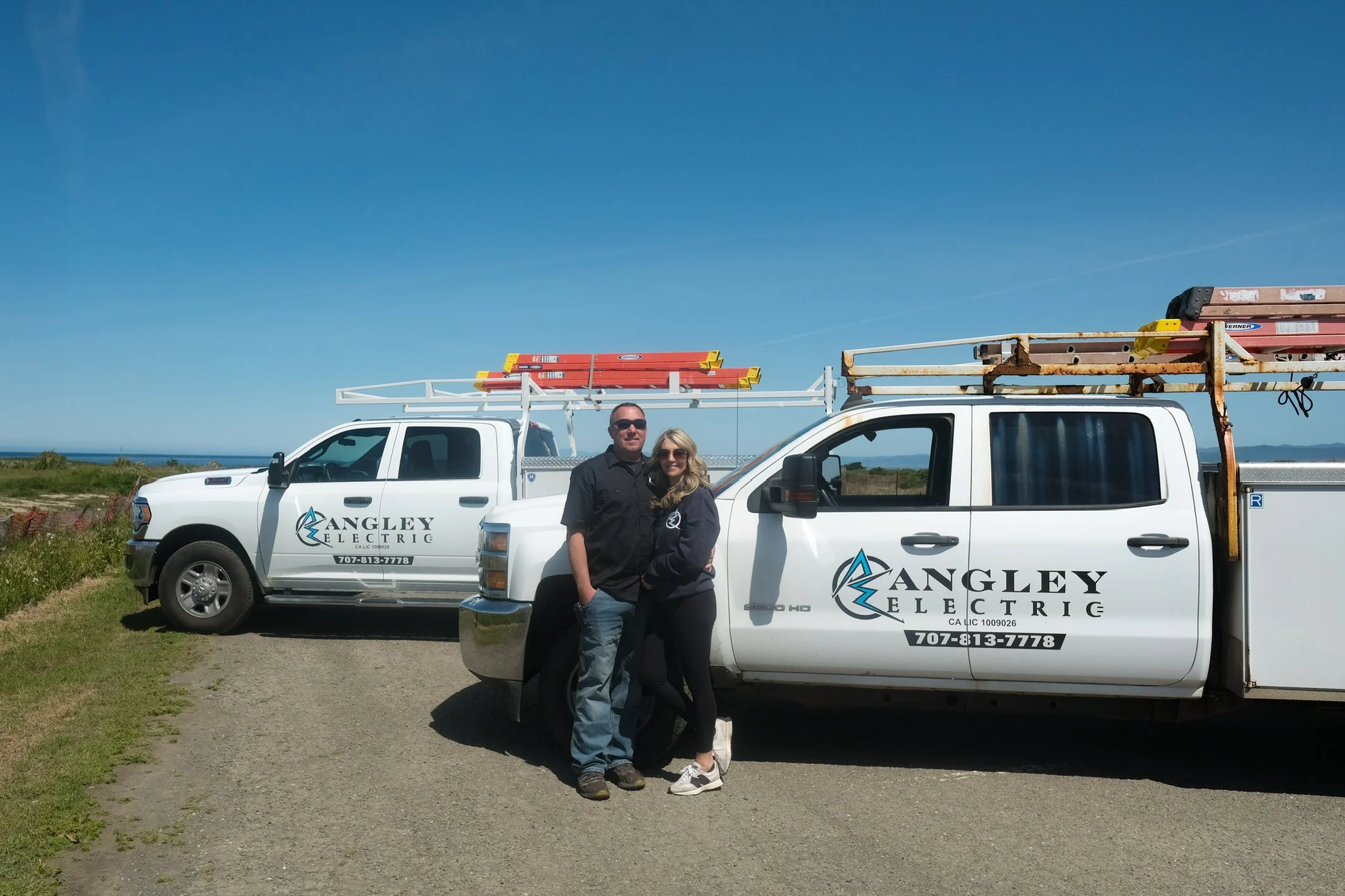 A man and woman standing in front of two white trucks with 'Angley Electric' logos, parked on a grassy area under a clear blue sky, with construction equipment on the trucks' roofs.
