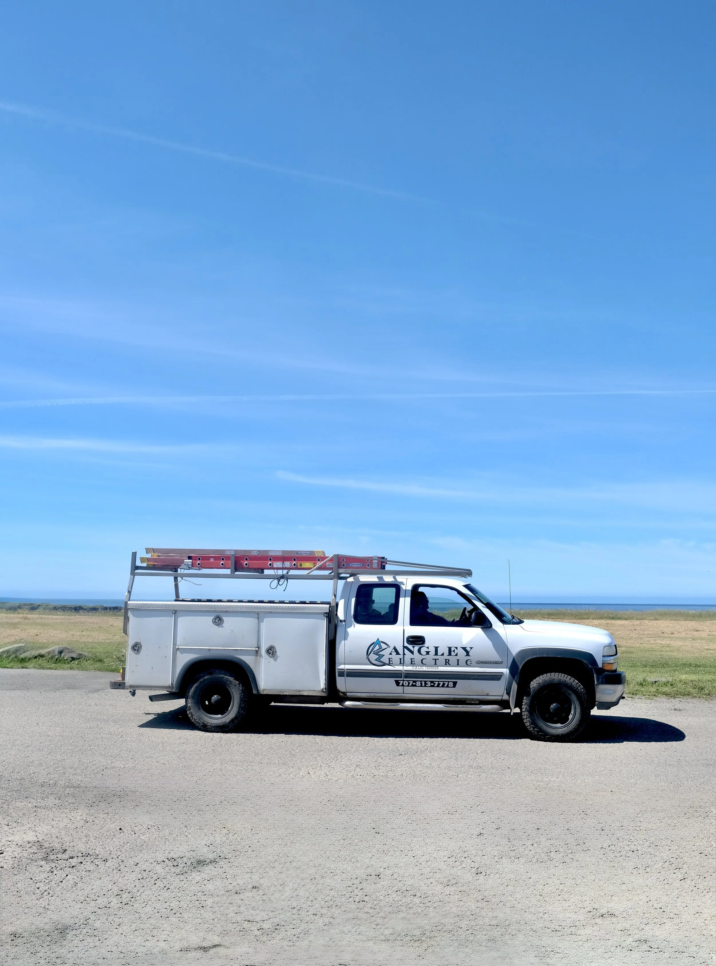 A white utility truck with company logo and contact information parked on roadside, with a clear blue sky overhead and grassy area in the background.