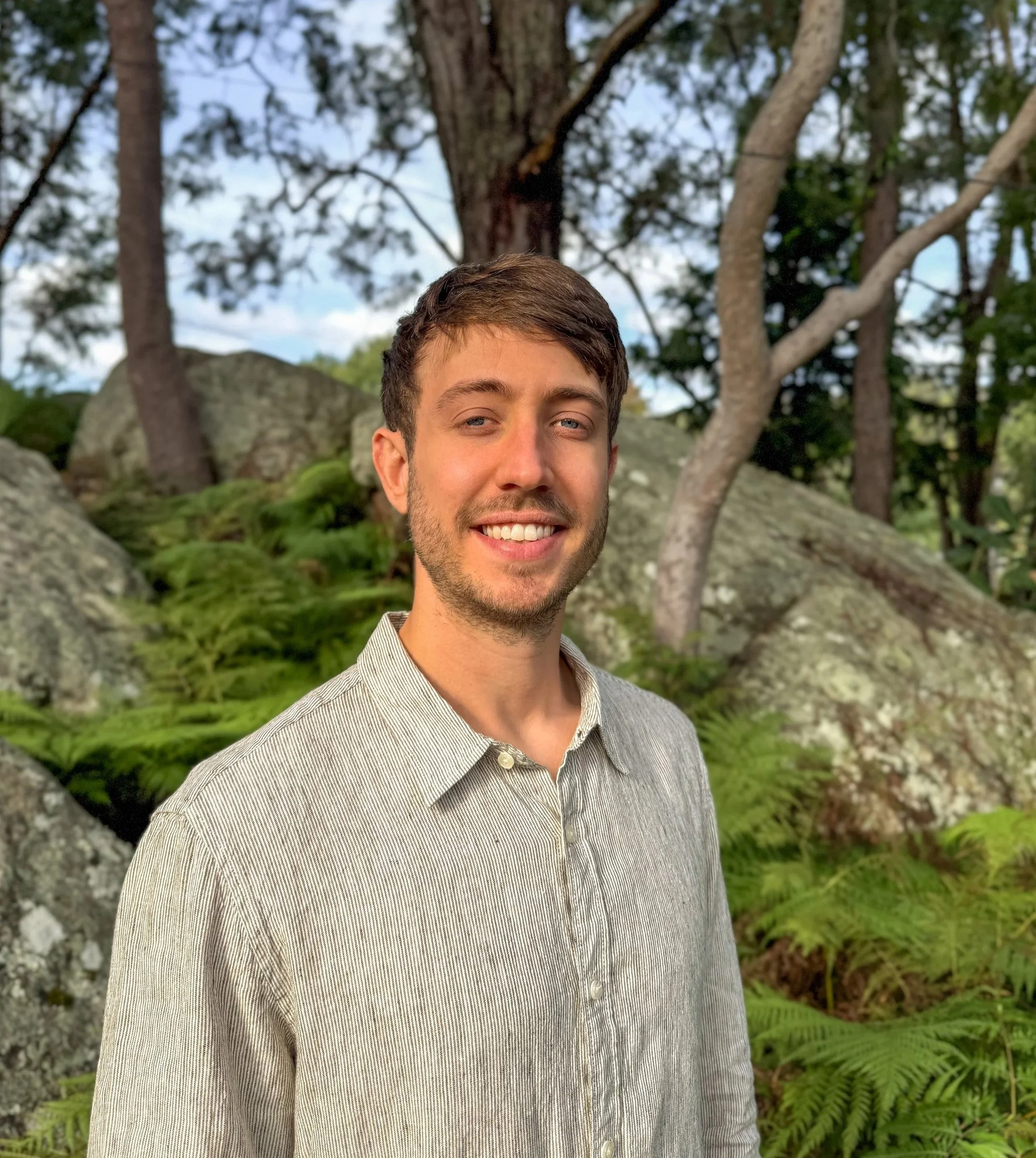 A young man with short brown hair, light beard, and a smile, standing outdoors in a forested area with rocks, green moss, and trees in the background.