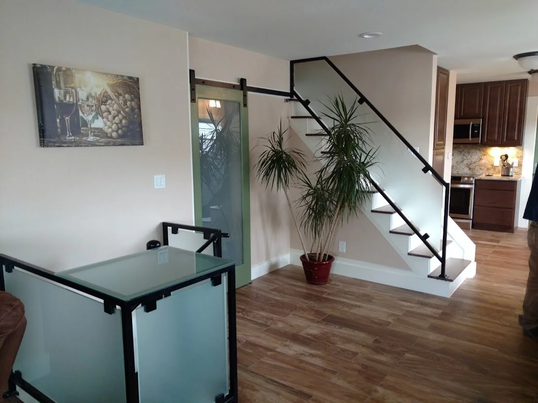 Living room with a staircase, large potted plant, glass-top table, artwork on the wall, and kitchen in the background.