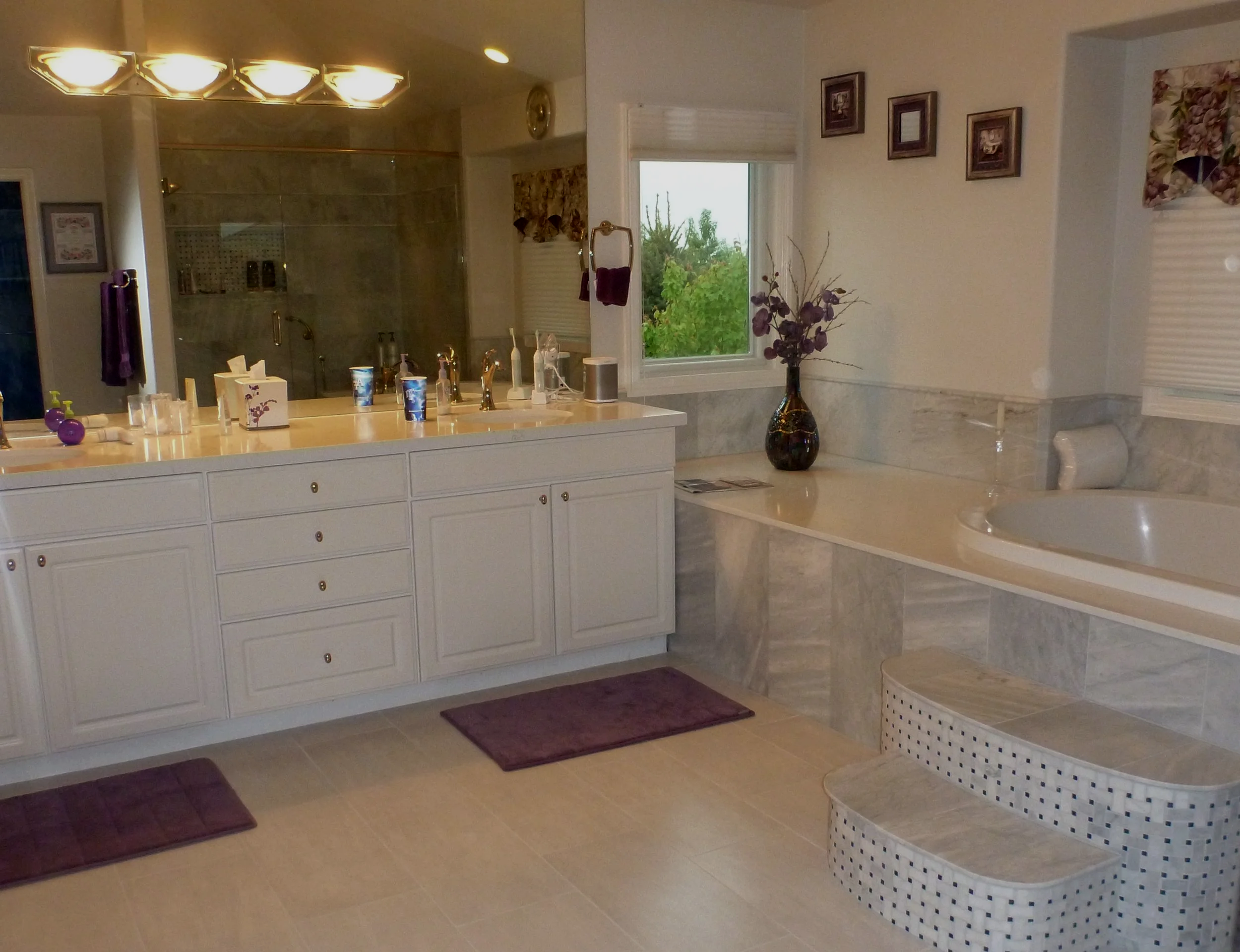 A bathroom with a large mirror over a white vanity, a bathtub with a tiled surround, a window with a view of greenery, framed pictures on the wall, a large vase with decorative flowers, and purple bath mats on the floor.