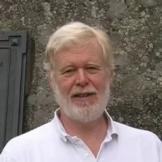A middle-aged man with white hair and a beard, wearing a white shirt, standing outdoors.
