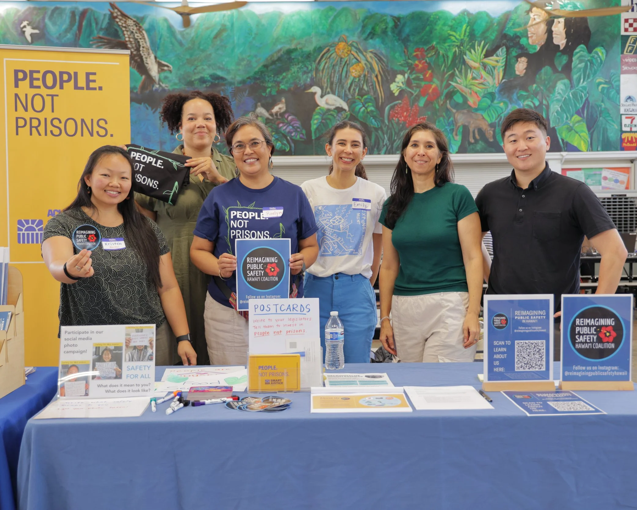 Six diverse individuals standing behind a table at an event promoting public safety and awareness, with signs and materials on the table, including a large yellow banner reading "People. Not Prisons."