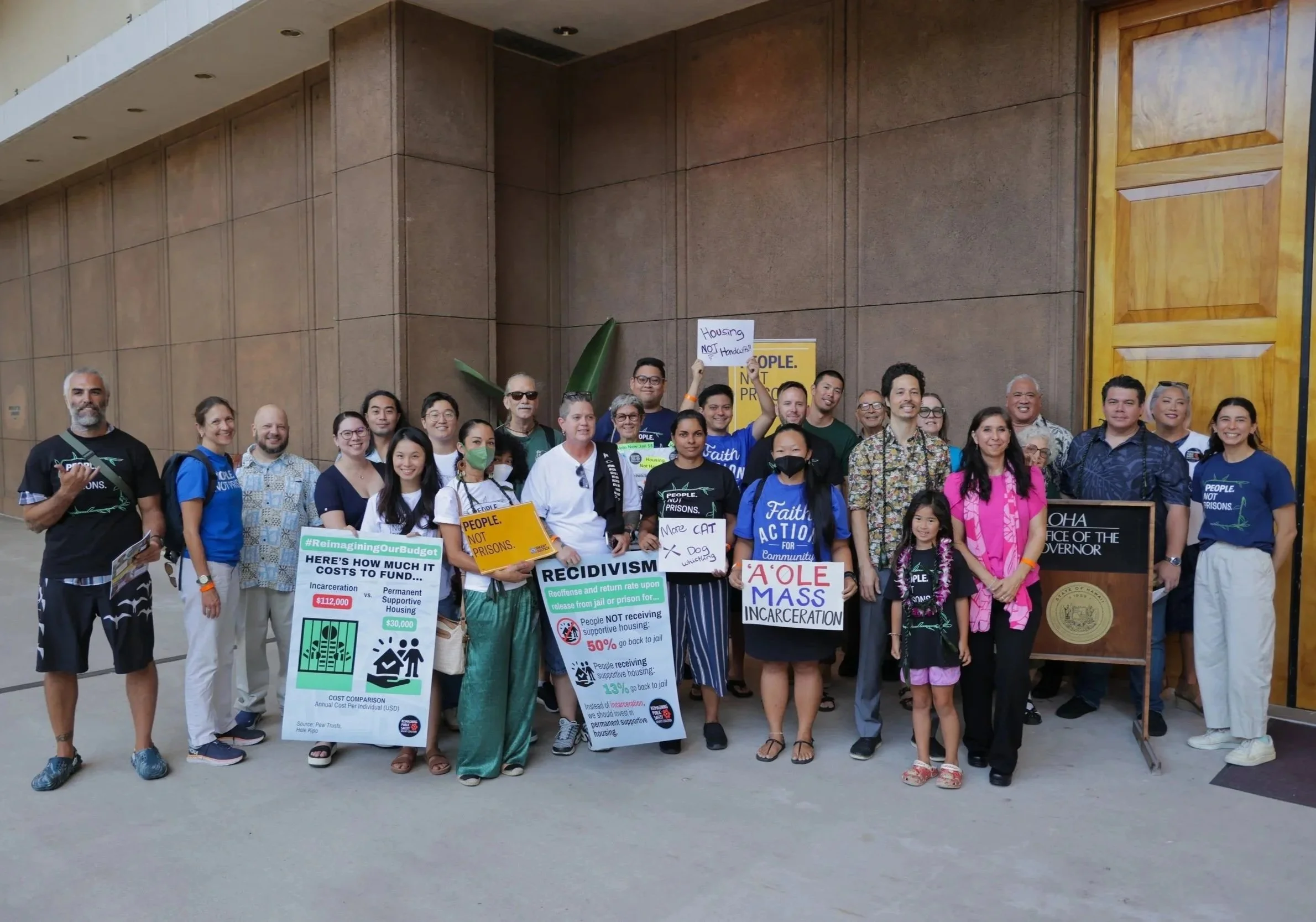 Group of people protesting outside a large building, holding signs and banners advocating against incarceration and housing issues, with some signs mentioning 'reimagining our budget' and 'A'OLE MASS incarceration'.