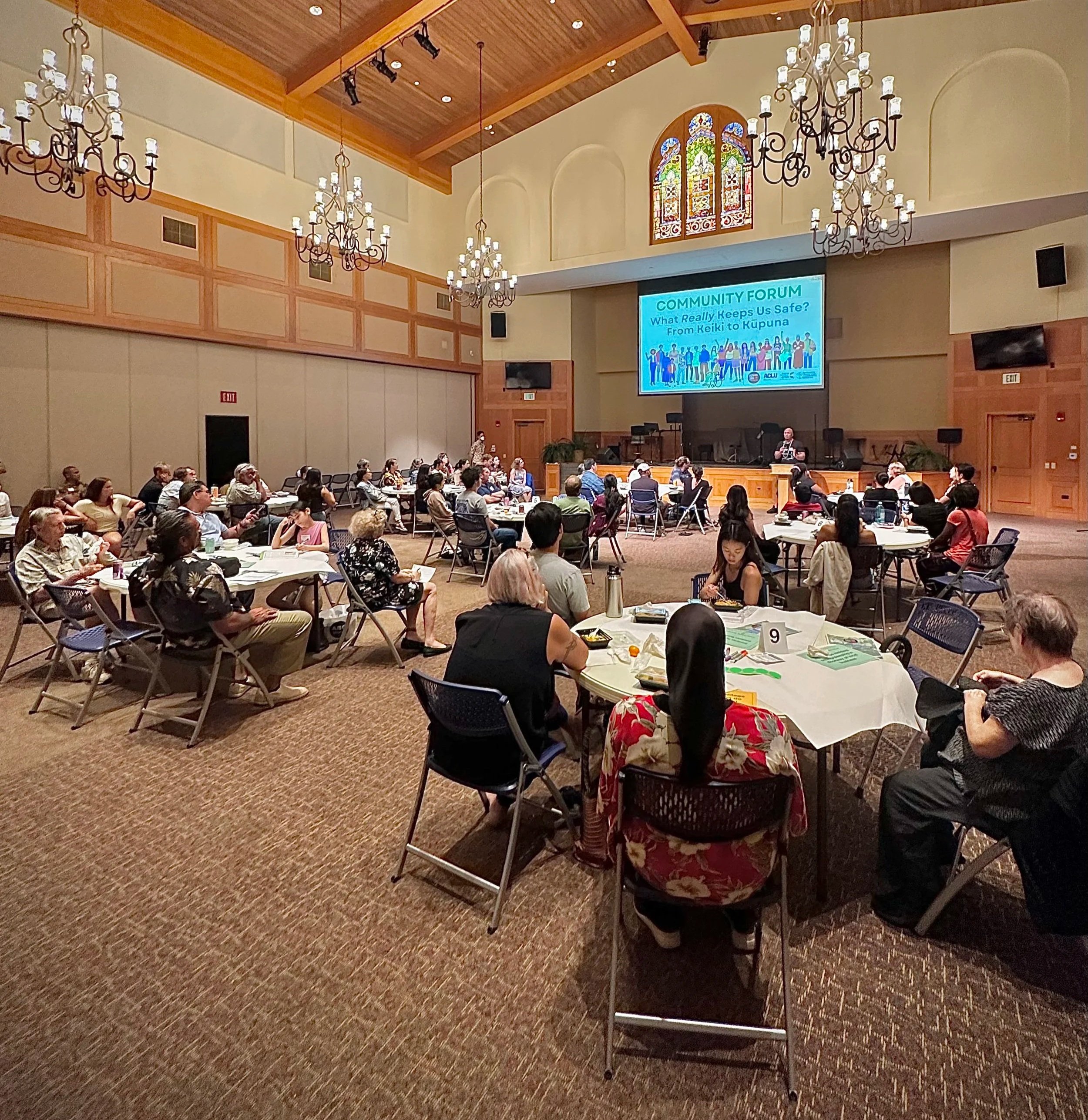 A large meeting room filled with round tables of people attending a community forum, with a stage at the front. There are chandeliers, wood-paneled walls, and stained glass windows. A presenter is speaking on the stage in front of a large screen displaying the forum's title.