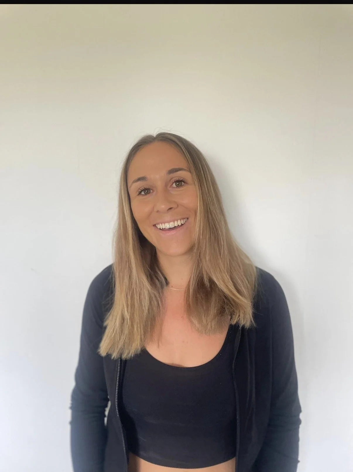 A woman with long light brown hair smiling and standing against a plain white wall, wearing a black zip-up jacket over a black crop top.