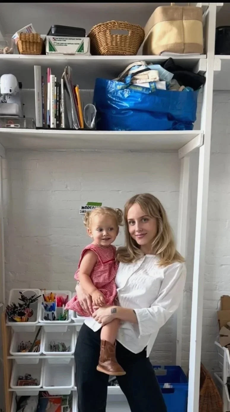 A woman holding a young girl in a room with shelves containing office supplies, books, and baskets.