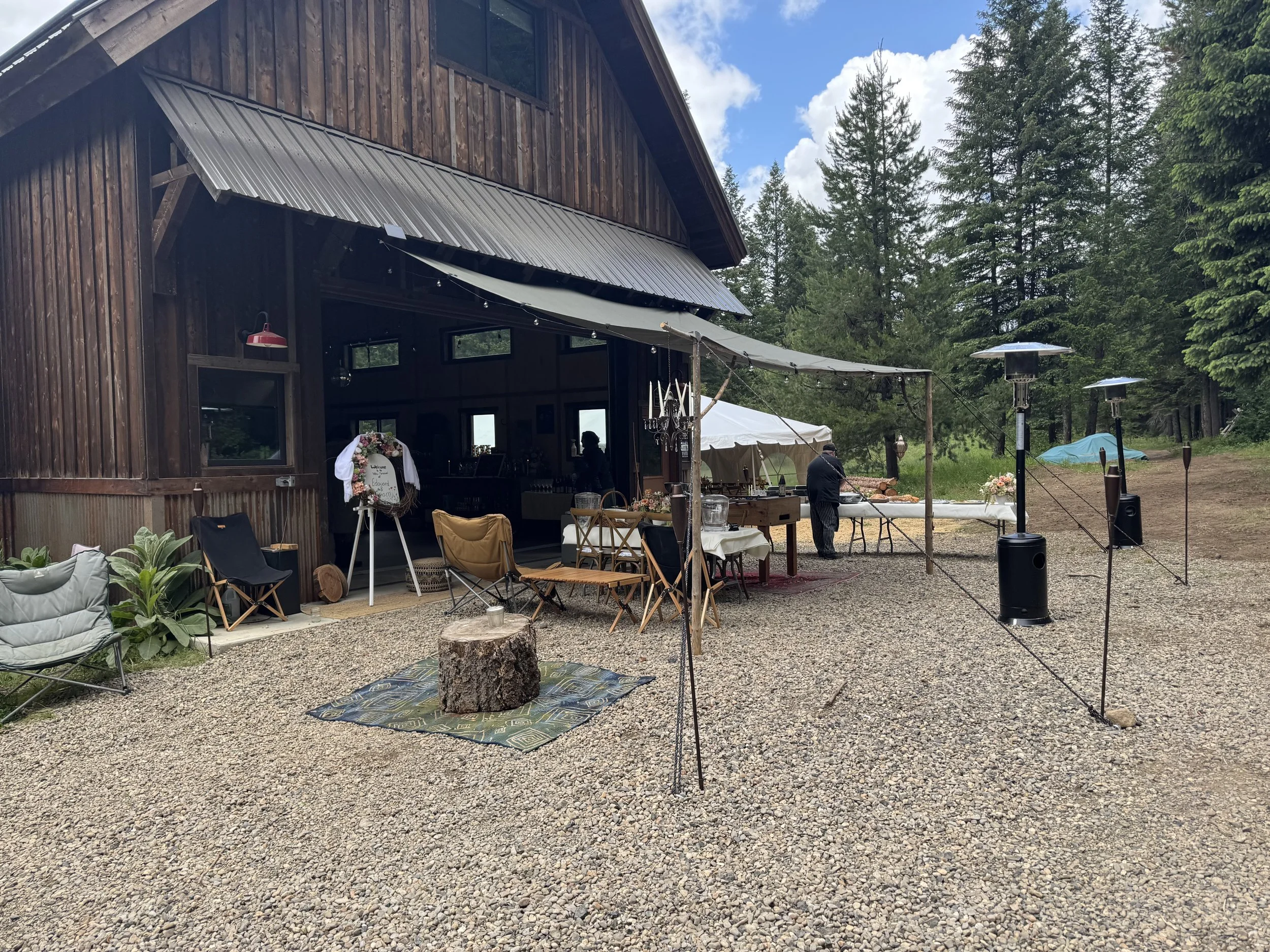 Outdoor wedding or celebration setup outside a wooden barn with chairs, tables, and a decorative flower wreath, in a forested area with trees and cloudy sky.