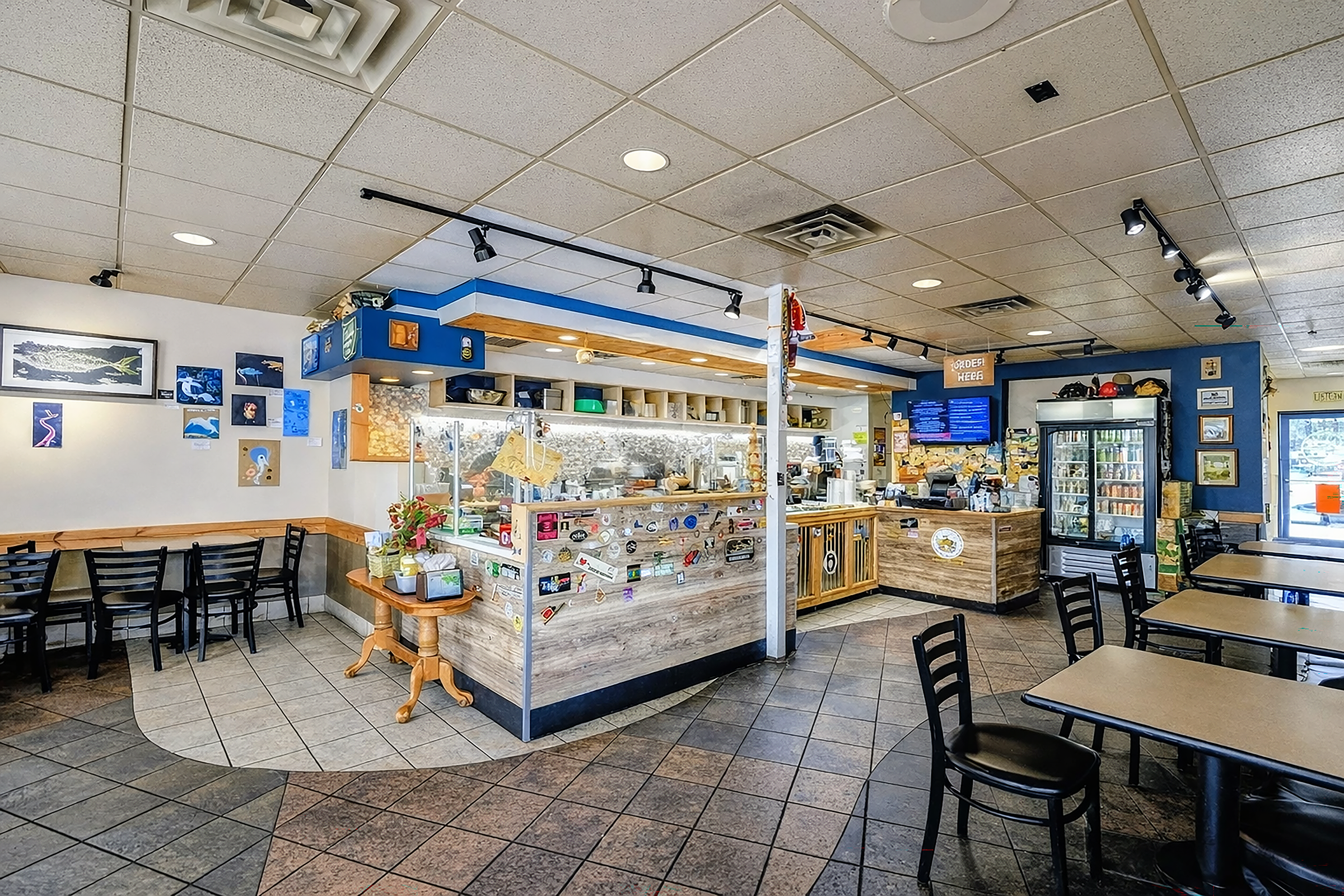 Interior of a casual restaurant with seating, a counter with a glass display case, a refrigerator, wall art, and digital menu boards.