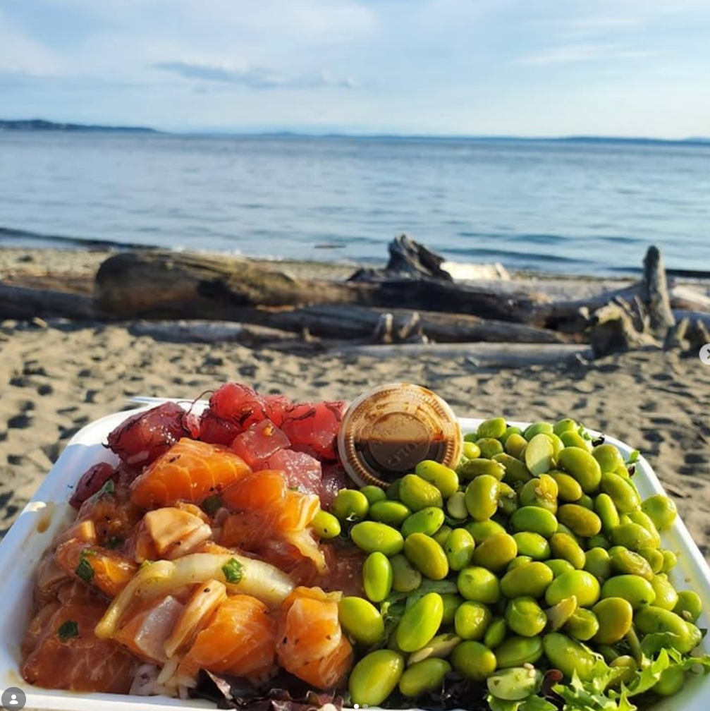 A plate of assorted raw fish and green beans on a beach with driftwood and the ocean in the background.