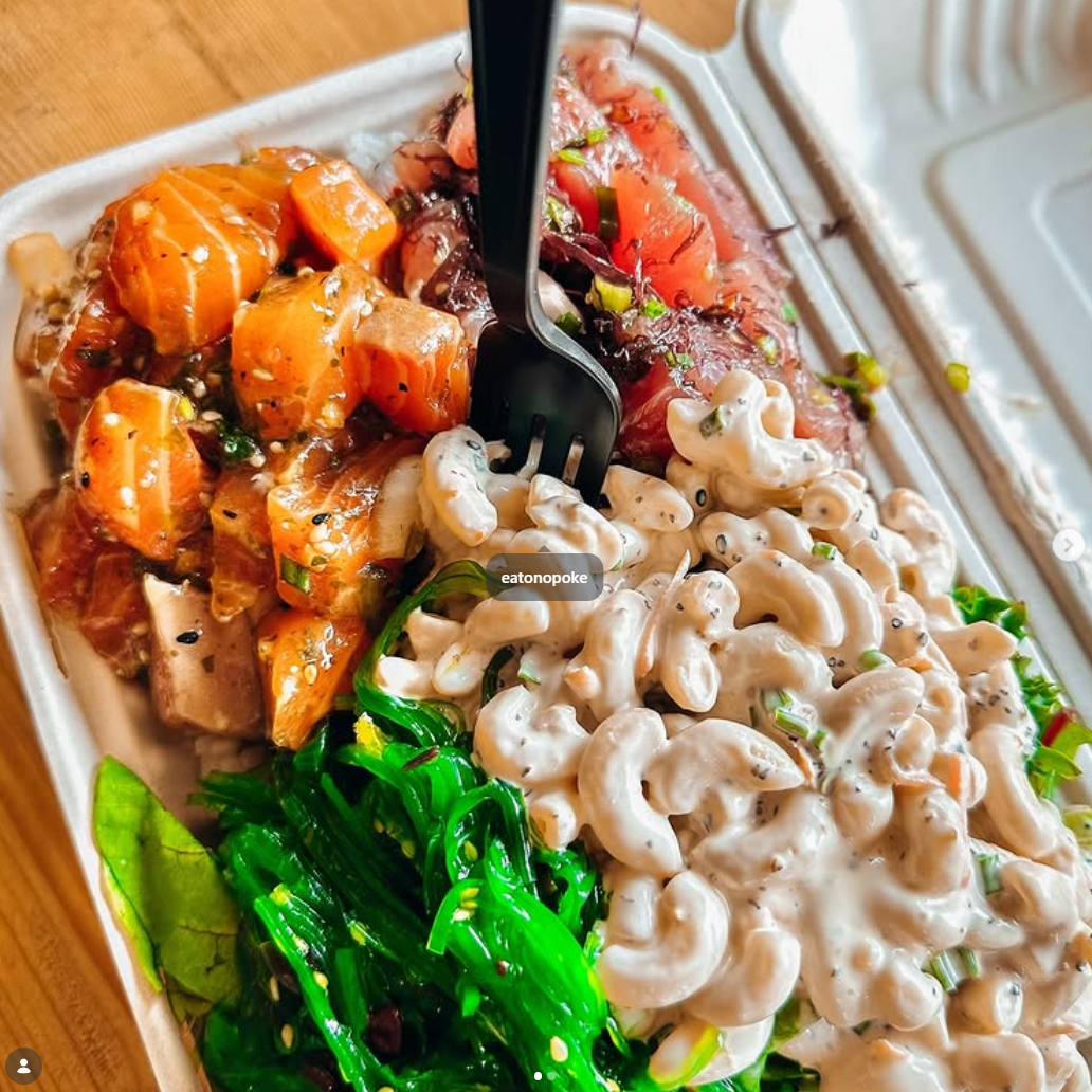 A takeout container filled with raw salmon, tofu, seaweed salad, and veggies, with a black fork inserted into the salmon.