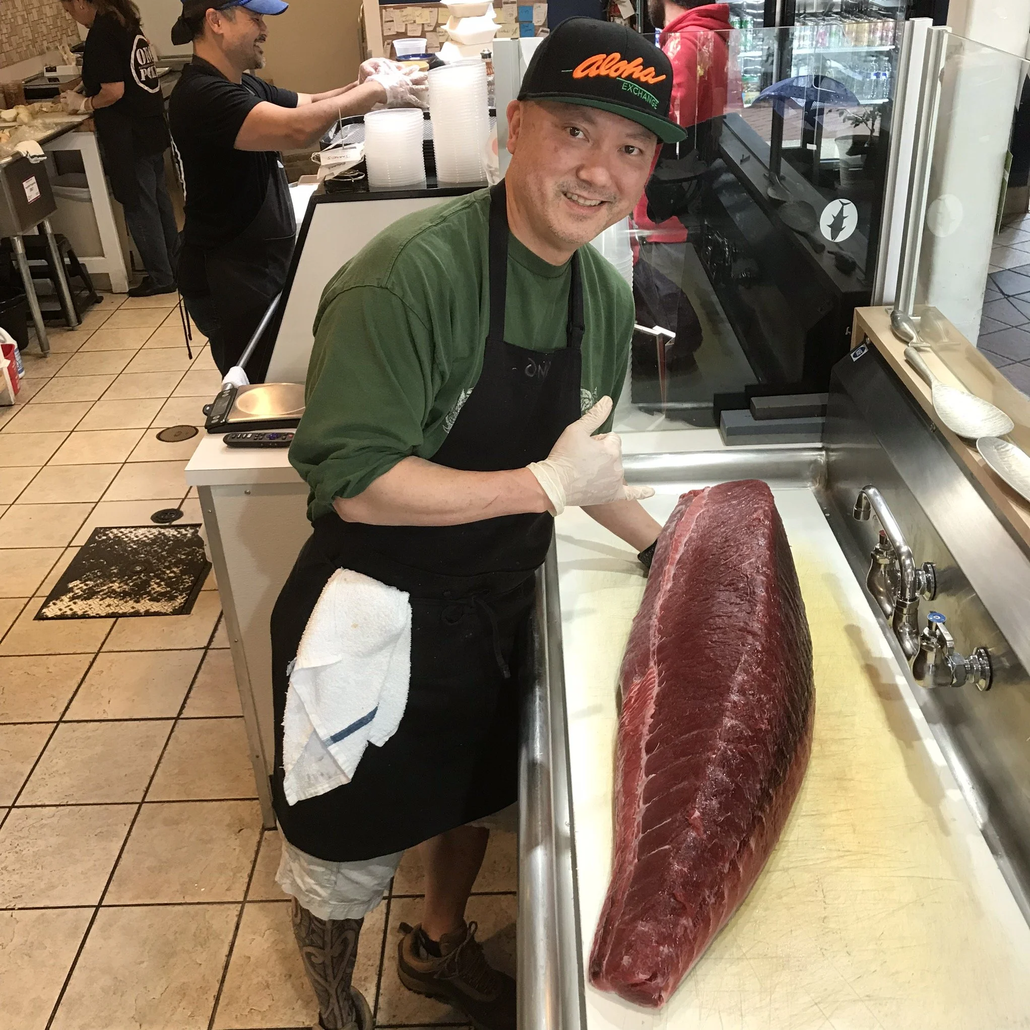 A man wearing a green shirt, black apron, and a black cap with orange and green lettering is standing at a meat counter with a large cut of raw beef. He is smiling and giving a thumbs-up gesture. There are other people working in the background in a busy kitchen or butcher shop.