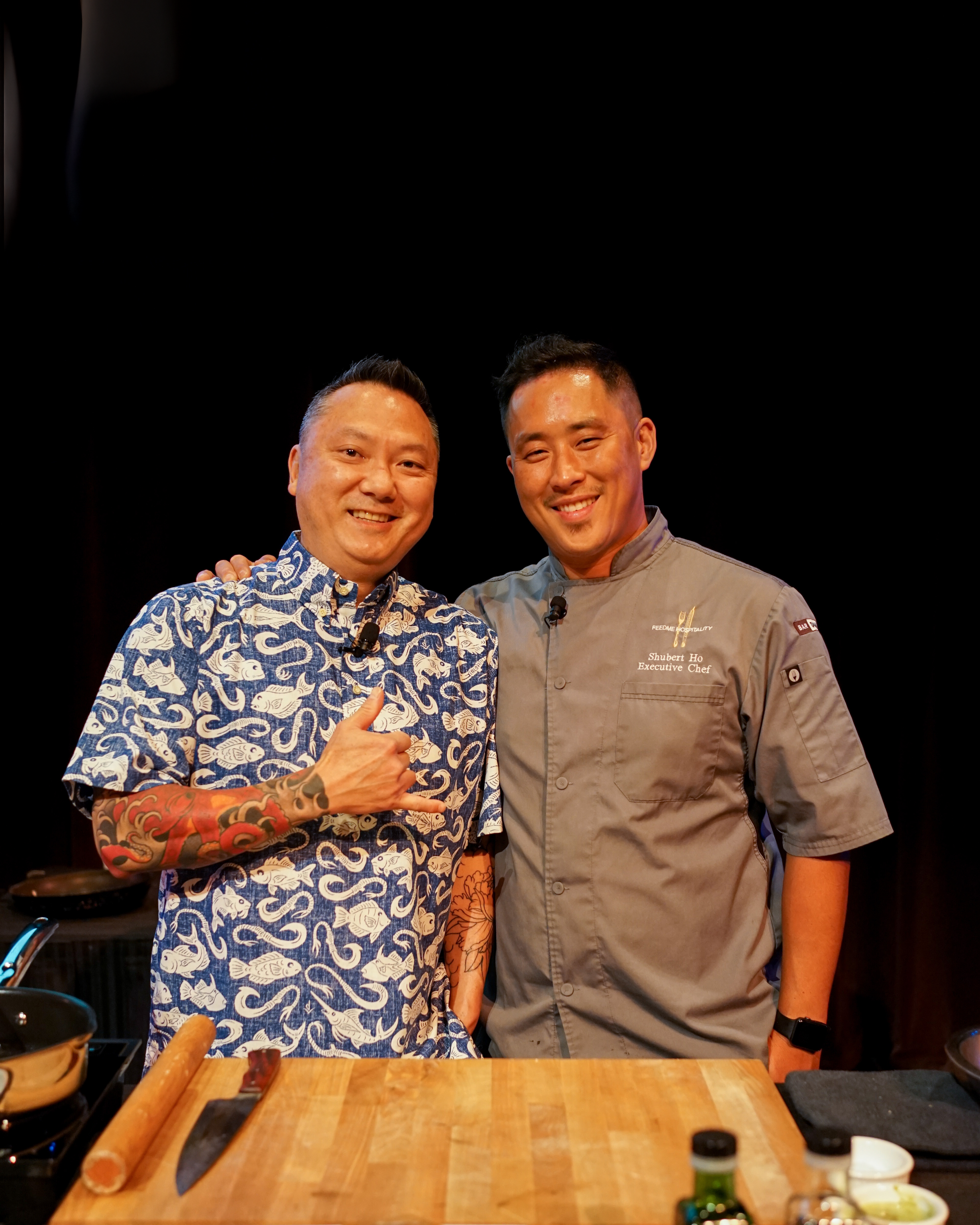 Two men standing close together in a kitchen or cooking studio, smiling at the camera. One is wearing a blue patterned shirt with a tattooed arm, making a hand gesture, and the other is wearing a gray chef's coat with a name tag reading 'Shubert Ho, Executive Chef'.