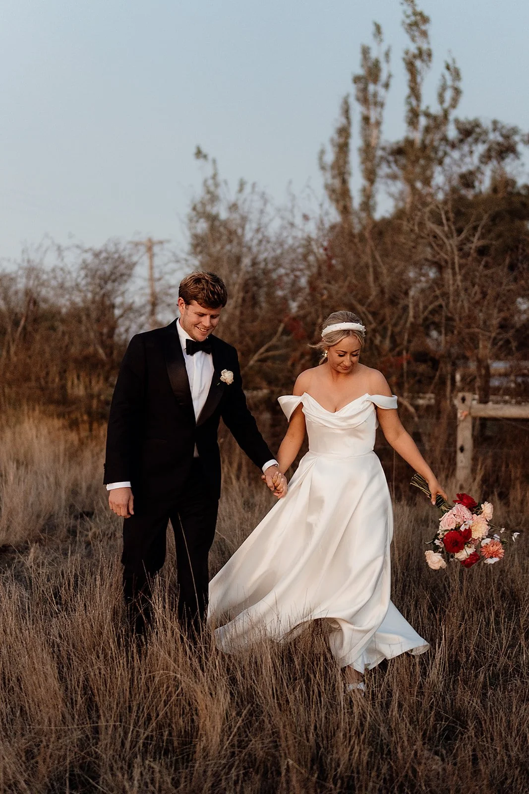 Bride and groom walking hand in hand through a grassy field during sunset, with the bride holding a bouquet of red and pink flowers, both smiling.