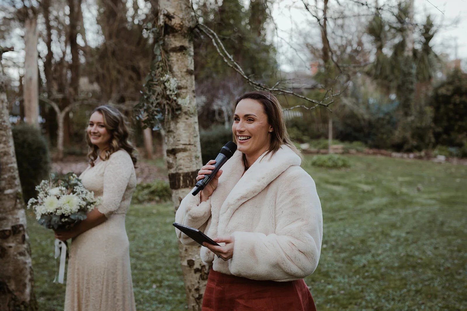 A woman in a light-colored coat holding a microphone and reading from a device, smiling outdoors with trees in the background during a wedding ceremony. The bride in a white dress, holding a bouquet, stands nearby.