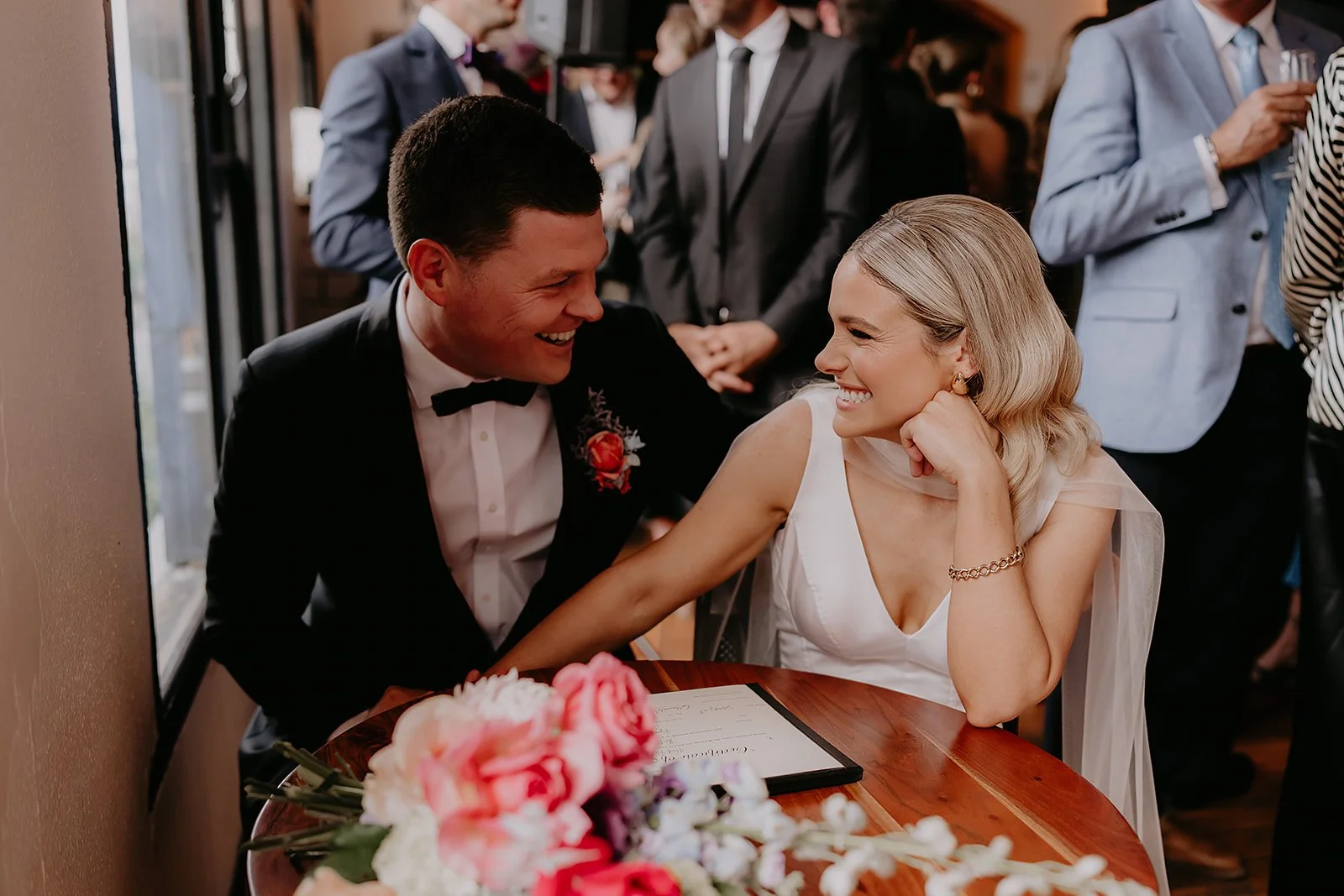 A happy couple, dressed in wedding attire, sitting at a table with a floral centerpiece, laughing and holding hands at their wedding reception.