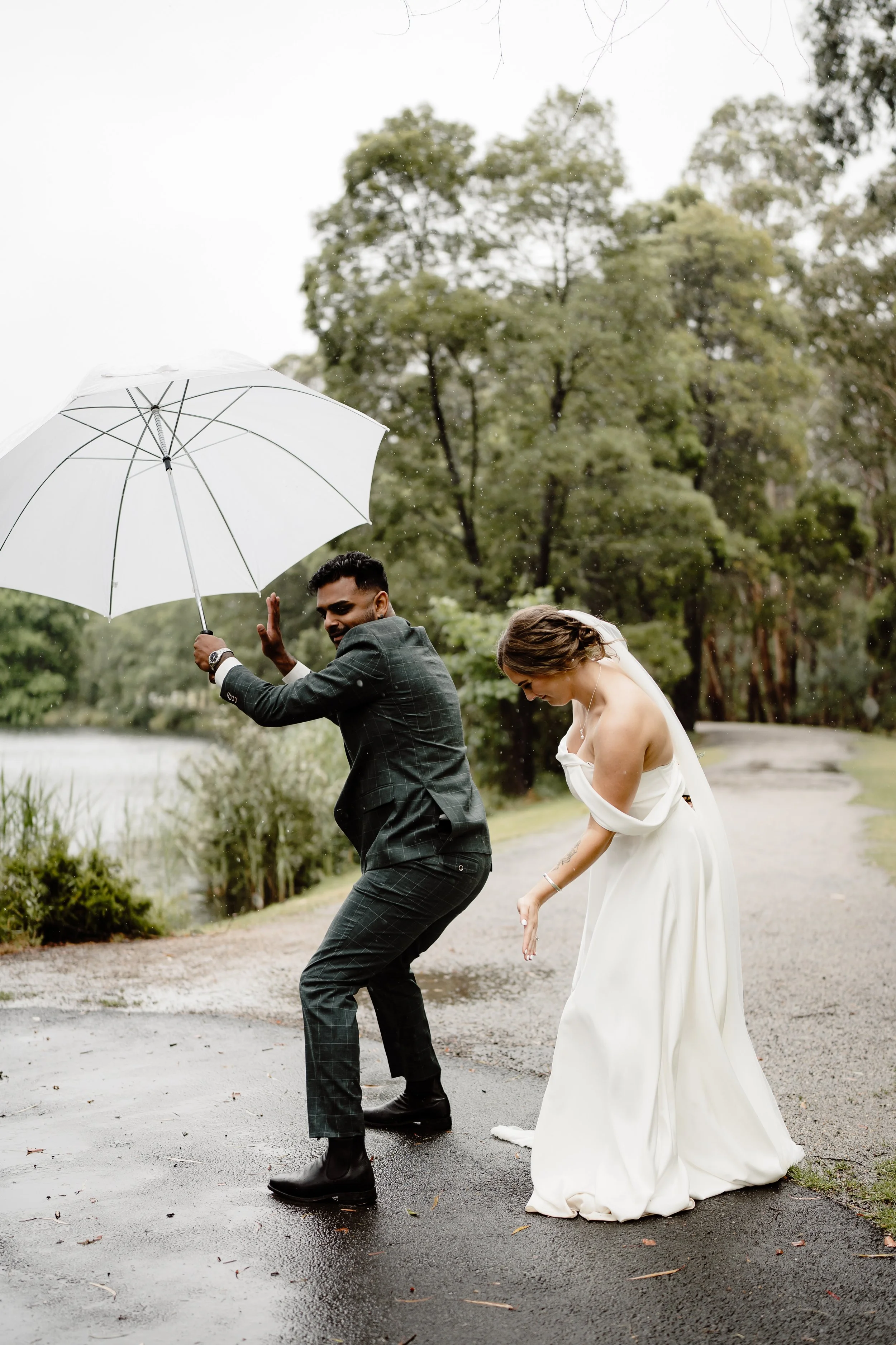A bride and groom holding an umbrella on a rainy day by a lake, with the groom dancing and the bride bowing.