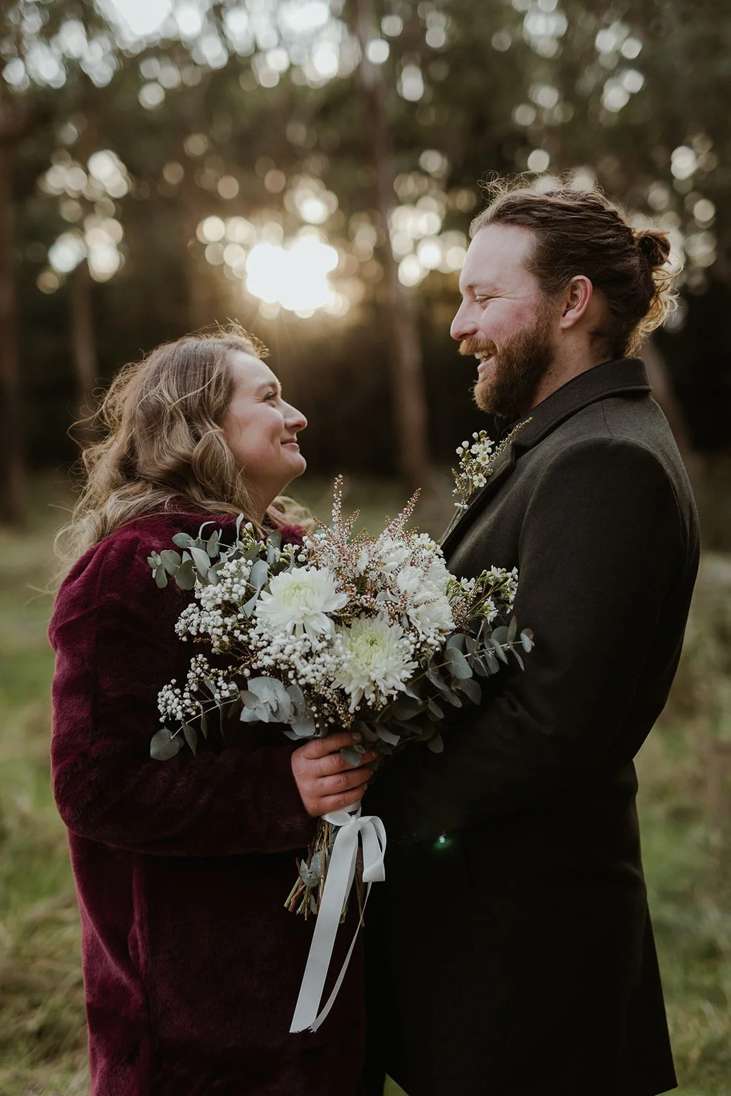 A woman and man standing face to face outdoors at sunset, holding a bouquet of white flowers and greenery, smiling at each other.
