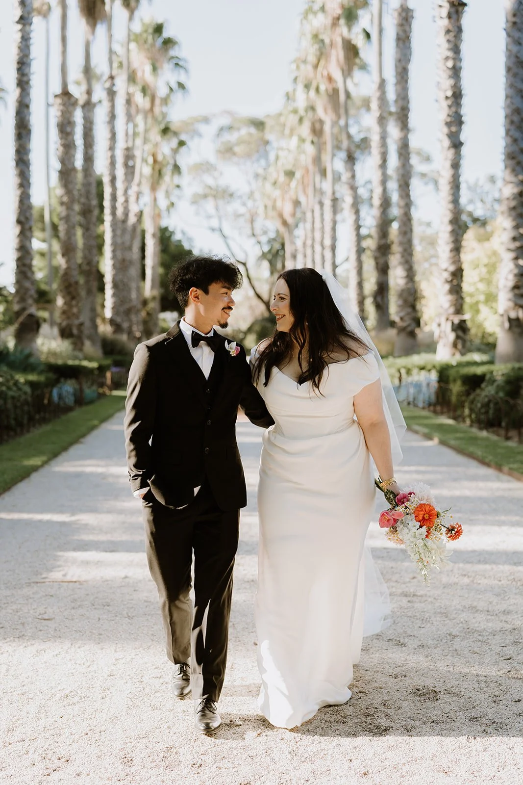 A bride and groom in wedding attire walk together outdoors, smiling and looking at each other, in a garden with tall palm trees and sunlight.