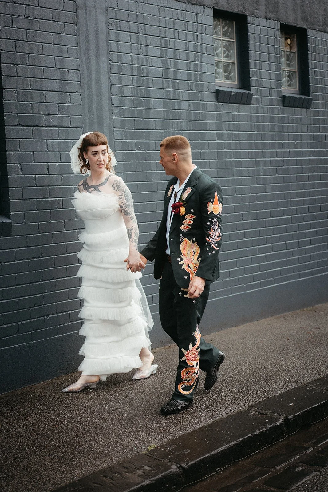 A bride and groom holding hands and walking on a wet sidewalk against a dark brick wall, dressed in wedding attire with the bride in a white ruffled dress and the groom in a black suit with colorful embroidery.