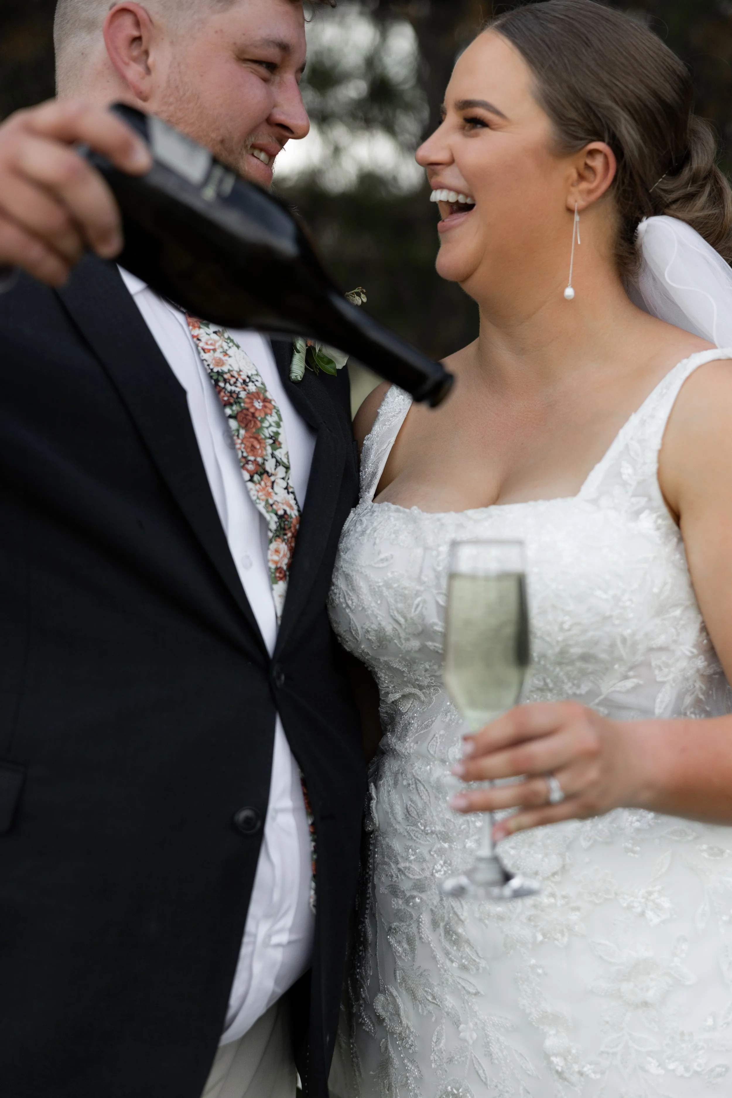 A bride and groom celebrating their wedding outdoors, smiling and holding glasses of champagne. The groom is holding a beer bottle.